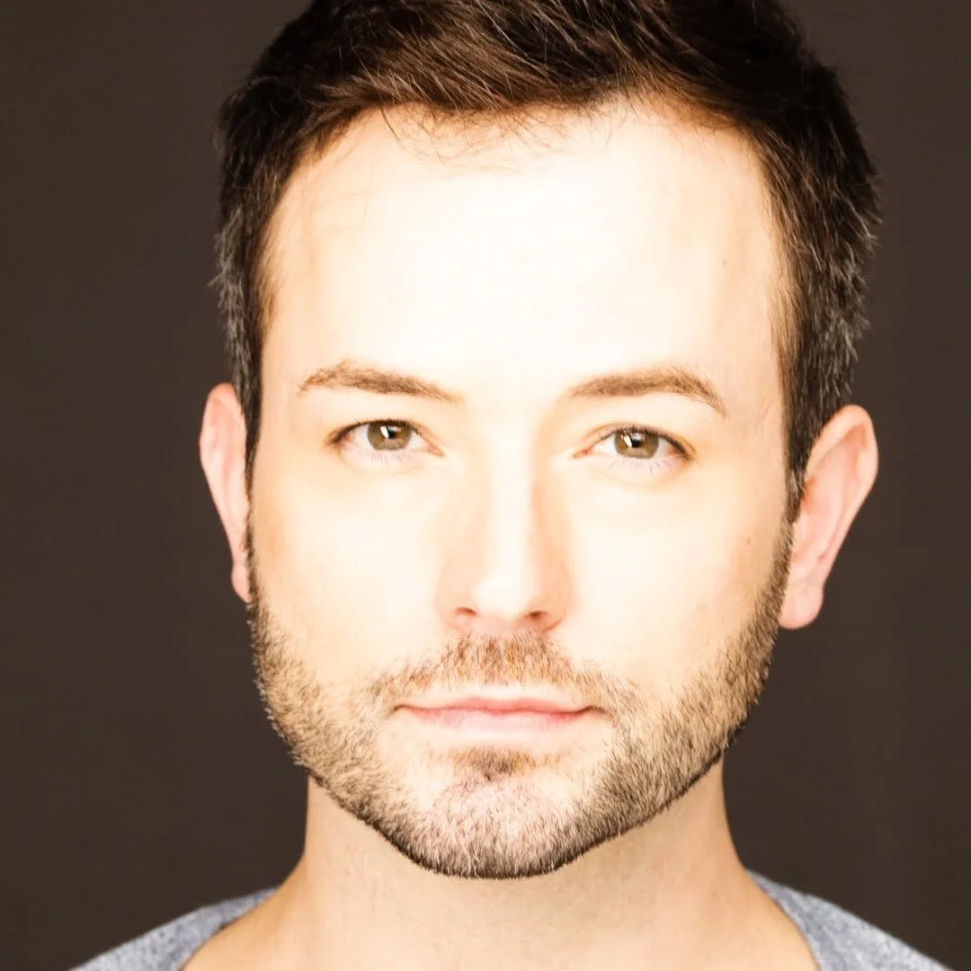 Close-up portrait of a man with dark hair, hazel eyes, and a trimmed beard against a dark background.