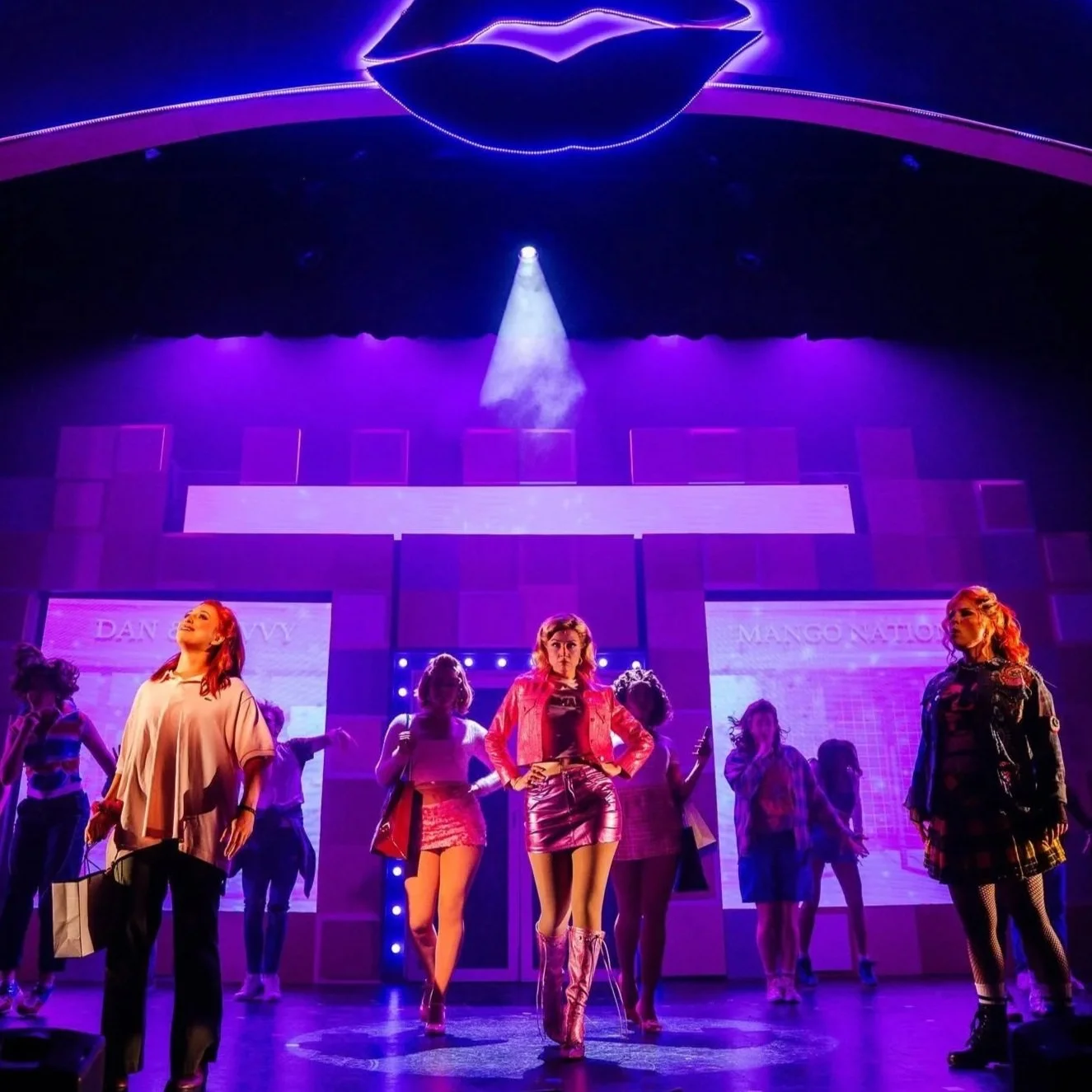 A group of young women with bright red and blonde hair performing on a stage with purple and blue lighting, multiple large screens in the background, and a lipstick-shaped light fixture hanging from the ceiling.