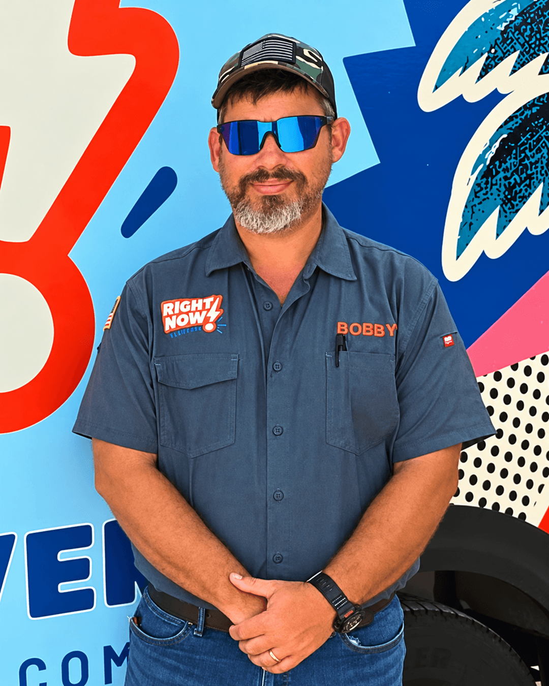 A man with a beard and sunglasses standing in front of a colorful, patterned background. He's wearing a dark blue work shirt with patches that say 'RIGHT NOW!' and 'BOBBY'.