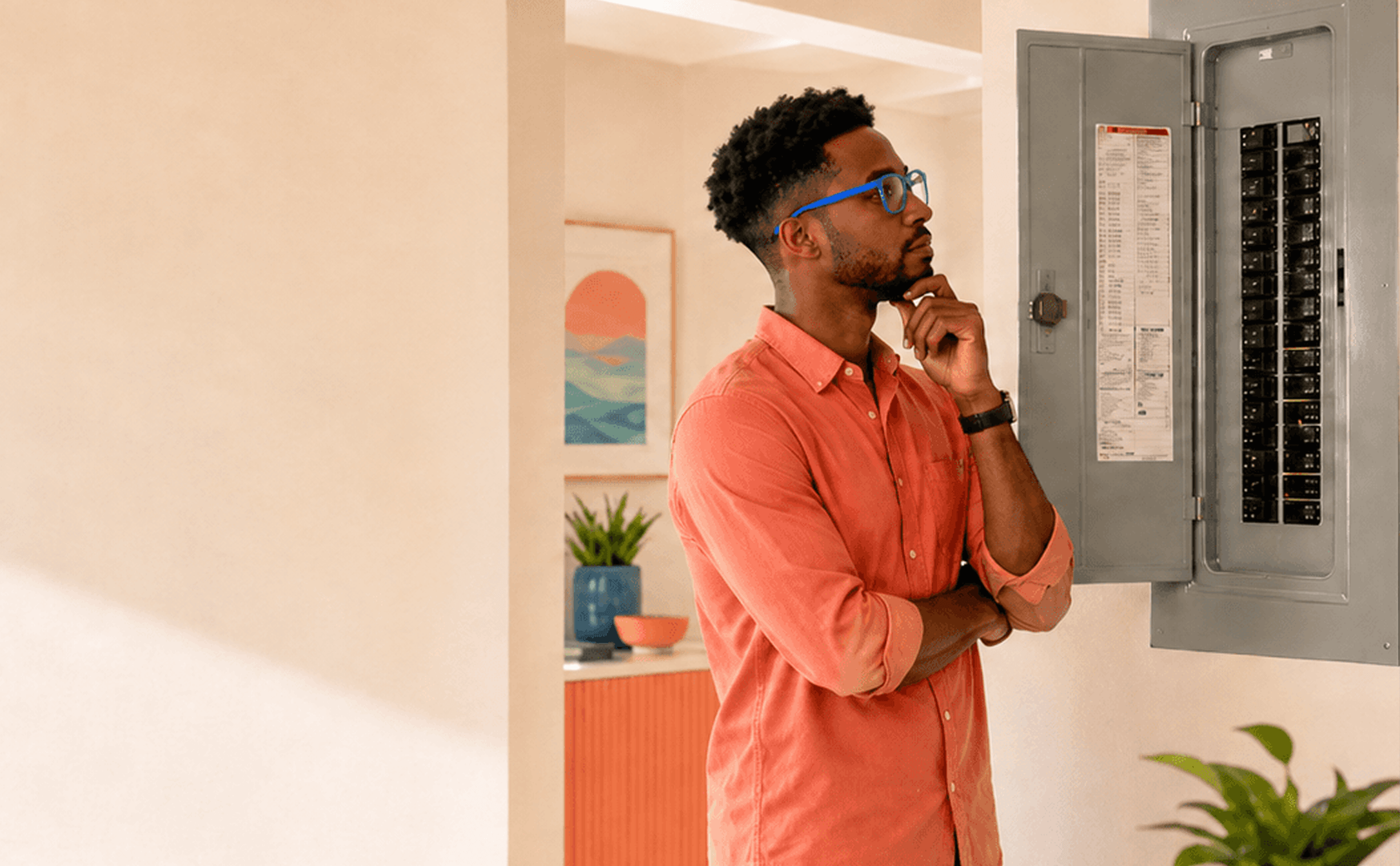 A man with curly hair, glasses, and a beard, wearing a salmon-colored shirt, is standing in front of an open electrical panel, thoughtfully looking at it with his hand on his chin in a modern, brightly decorated room.