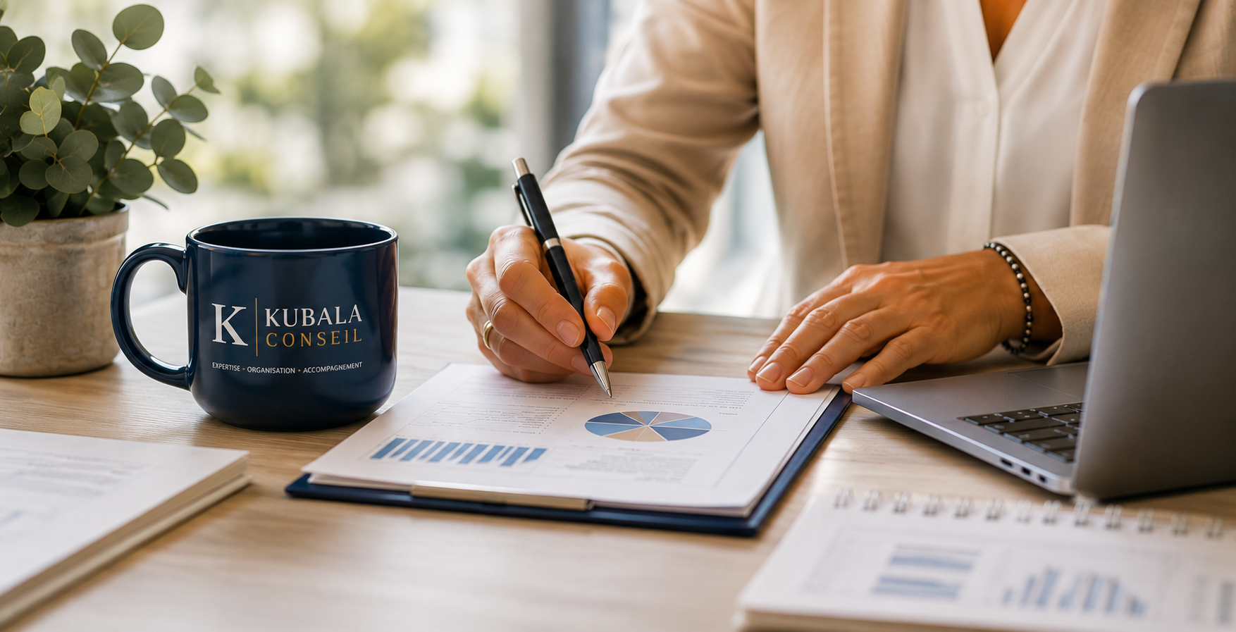 Une personne en costume beige rédige des documents avec des graphiques et des données sur une table en bois, à côté d'une tasse bleue portant la mention 'KUBALA CONSEIL' et un ordinateur portable, avec une plante verte en pot en arrière-plan.