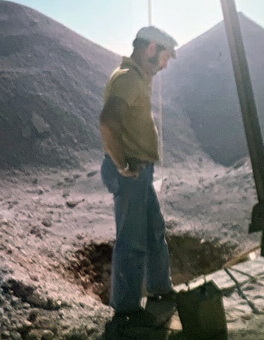 A man wearing a yellowish-green jacket and blue jeans stands on rocky terrain next to a large hole with his hands in his pockets. There are mountain slopes in the background.
