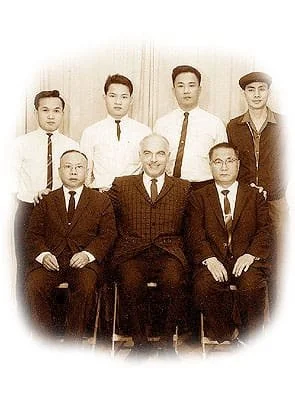 Group portrait of seven men, with four standing in back and three seated in front, dressed in formal suits and ties, in black and white.