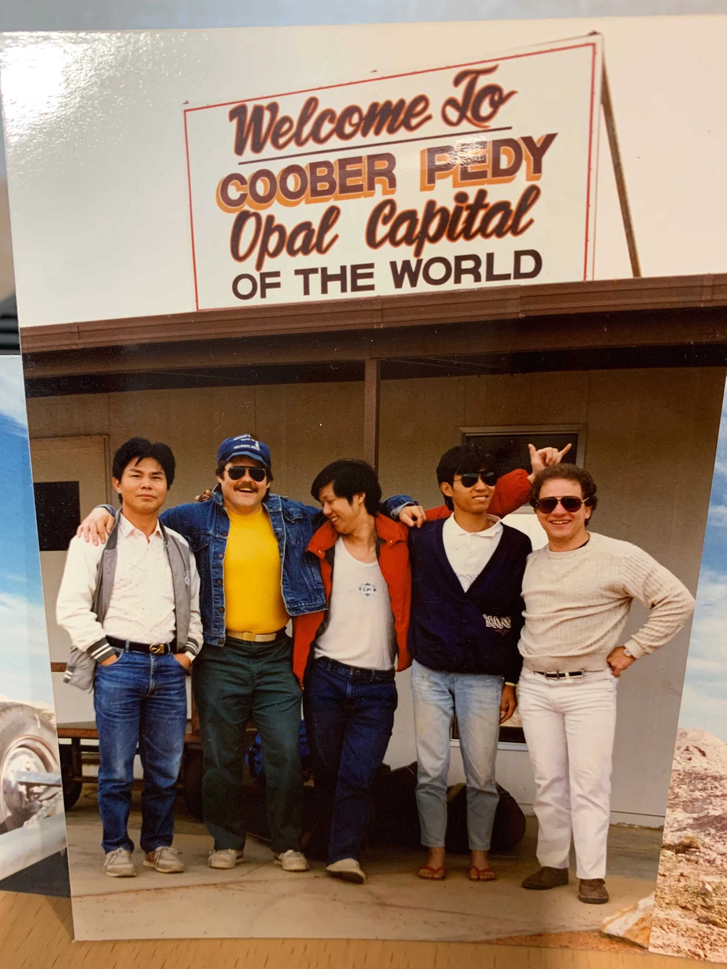 A group of six friends standing together in front of a sign that says "Welcome To Coober Pedy Opal Capital Of The World." The friends are smiling and wearing casual clothing, including sunglasses and jackets. The background includes parts of a building and some outdoor scenery.