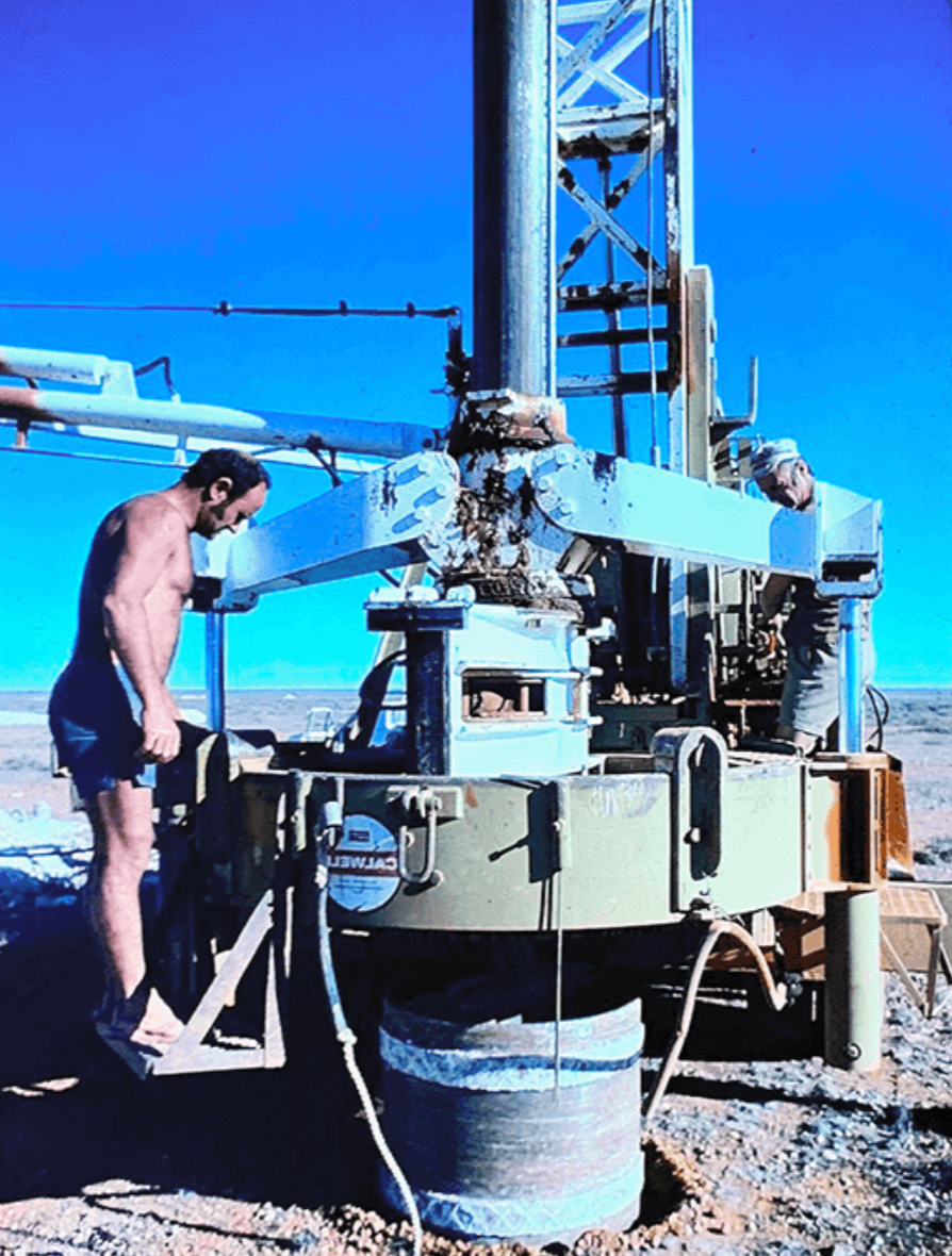 Two men operate a large oil or natural gas drilling rig in a dry, barren landscape under a clear blue sky.