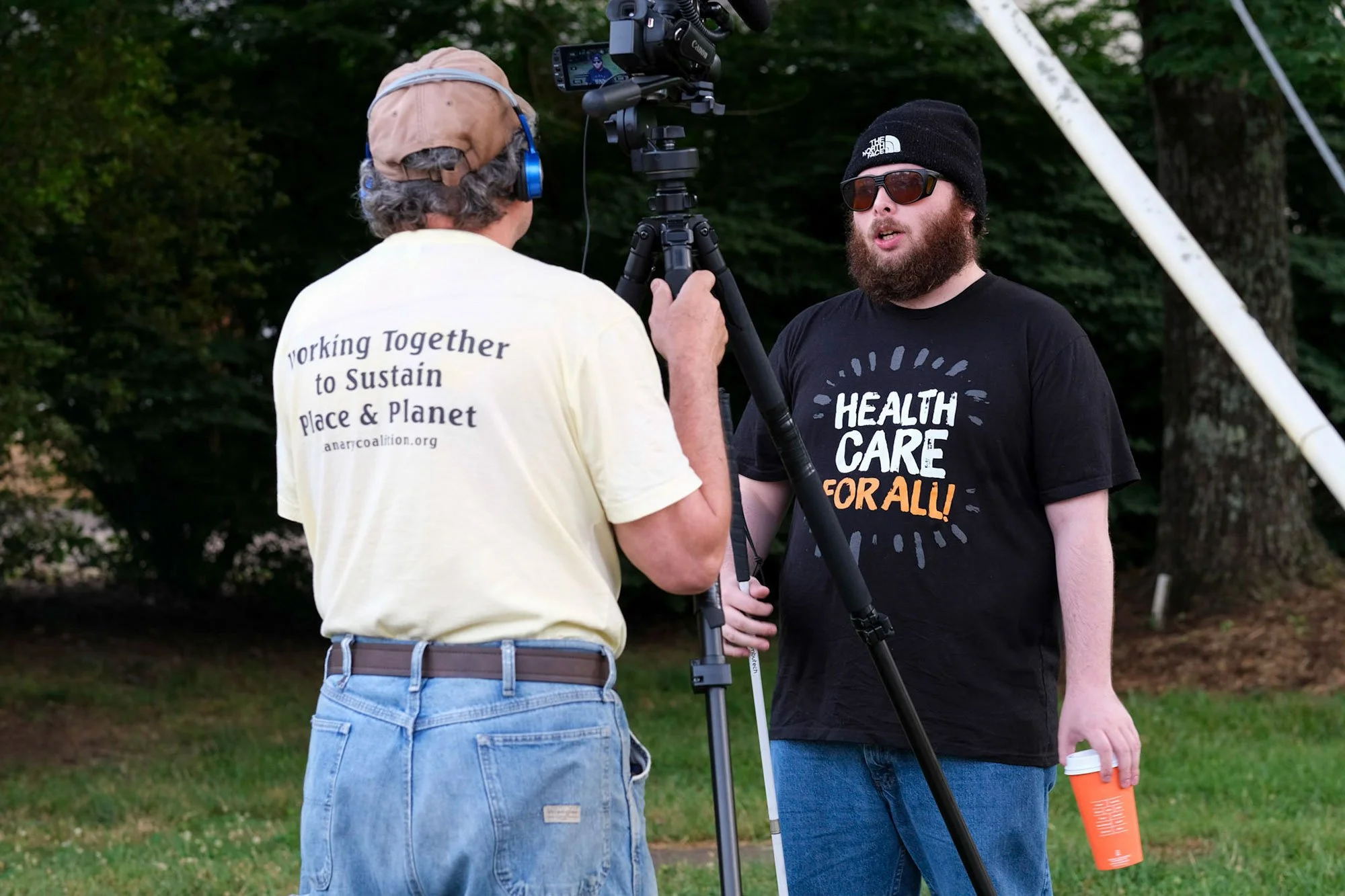 Man holding a camera in front of another man speaking who is wearing a shirt that says "health care for all"