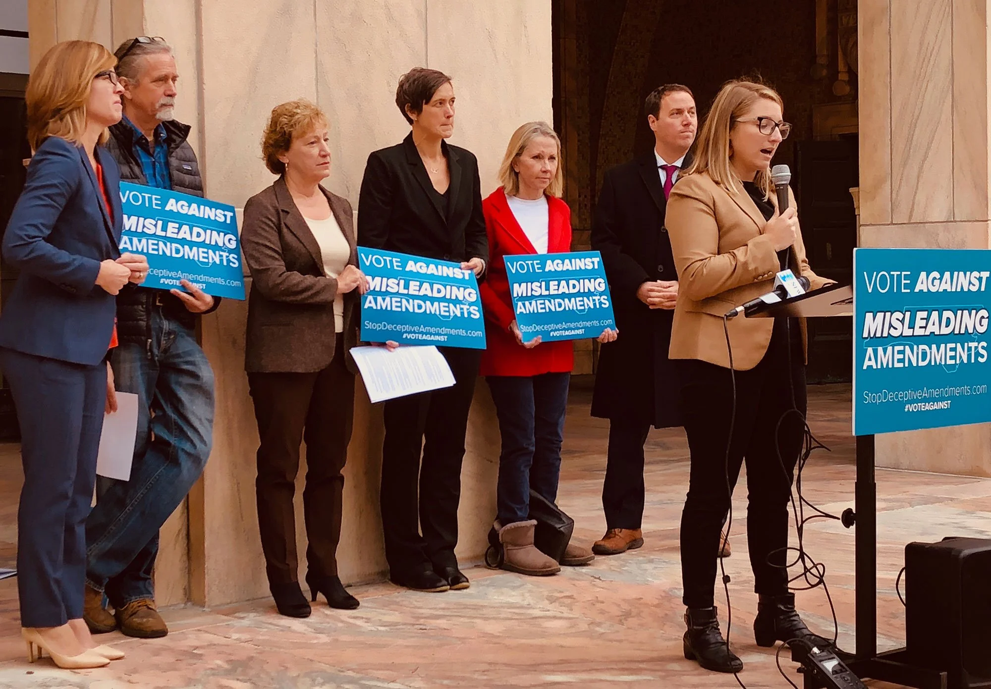 Woman speaking at a podium with people behind her holding signs that say "vote against misleading amendments"