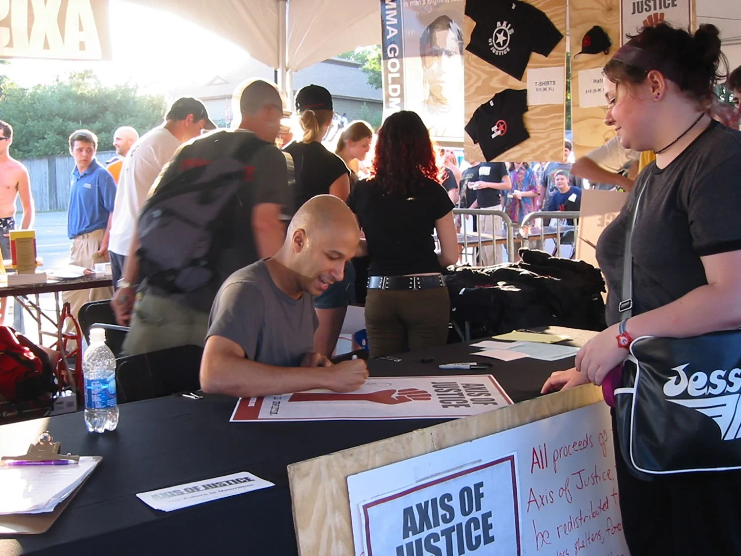 Man signing a poster that says "axis of justice" on a table at Lollapalooza, with a woman standing in front waiting for the poster