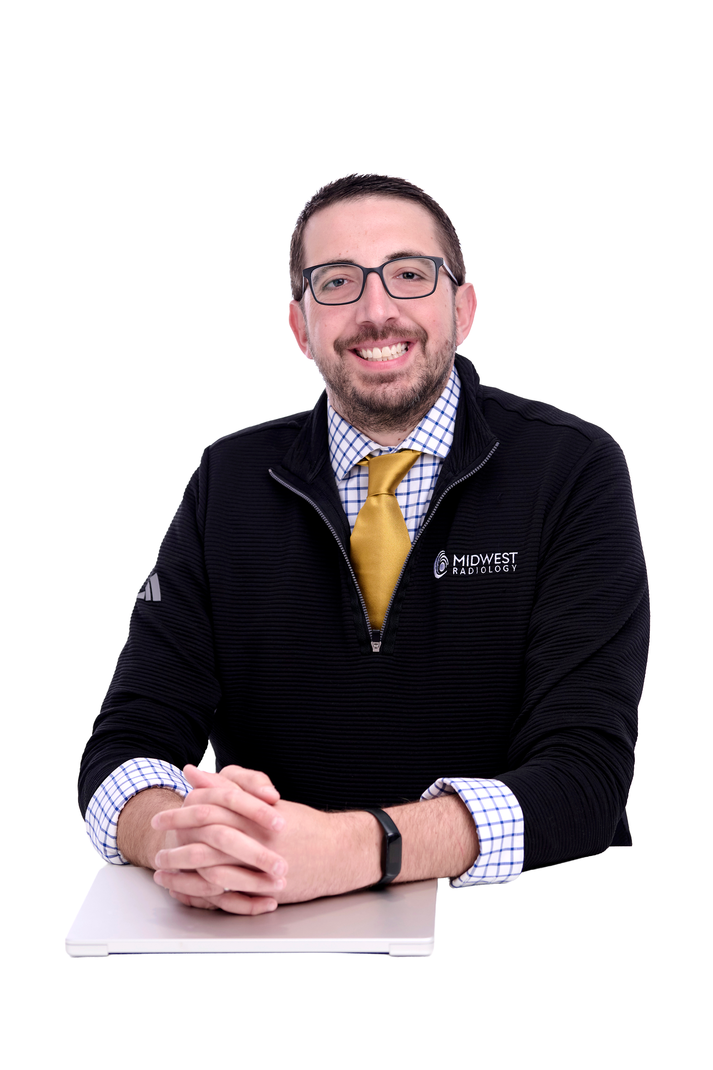 A young man with glasses, a beard, and a mustache smiling at the camera, sitting at a table with folded hands. He is wearing a black jacket with a logo that reads 'Midwest Radiology,' a blue and white checked shirt, a yellow tie, and a black wristwatch.