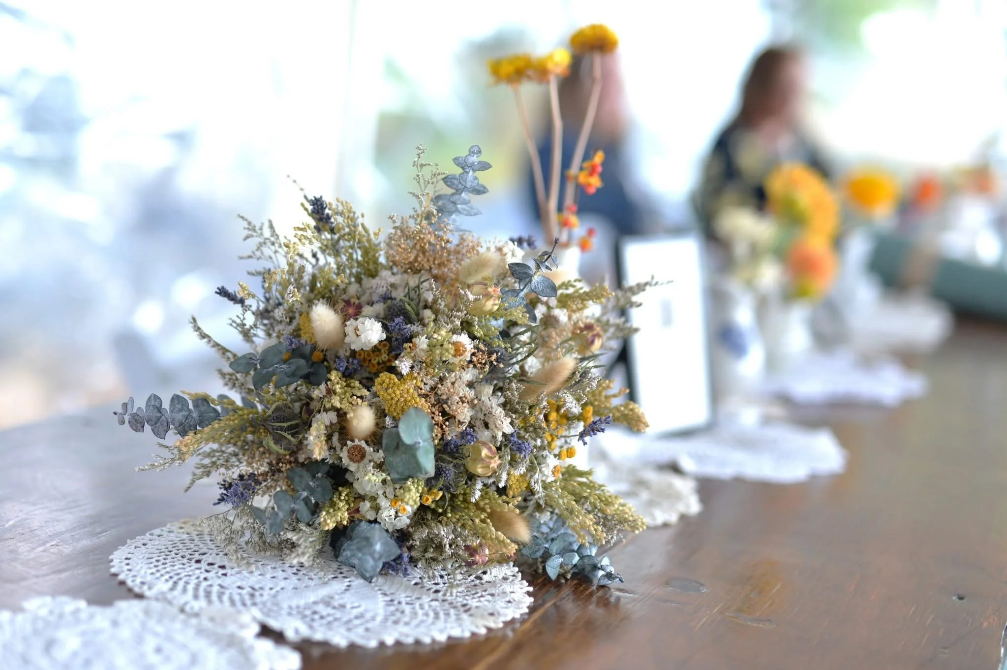 A bouquet of dried flowers with various colors and textures, placed on a wooden table with a lace doily underneath.