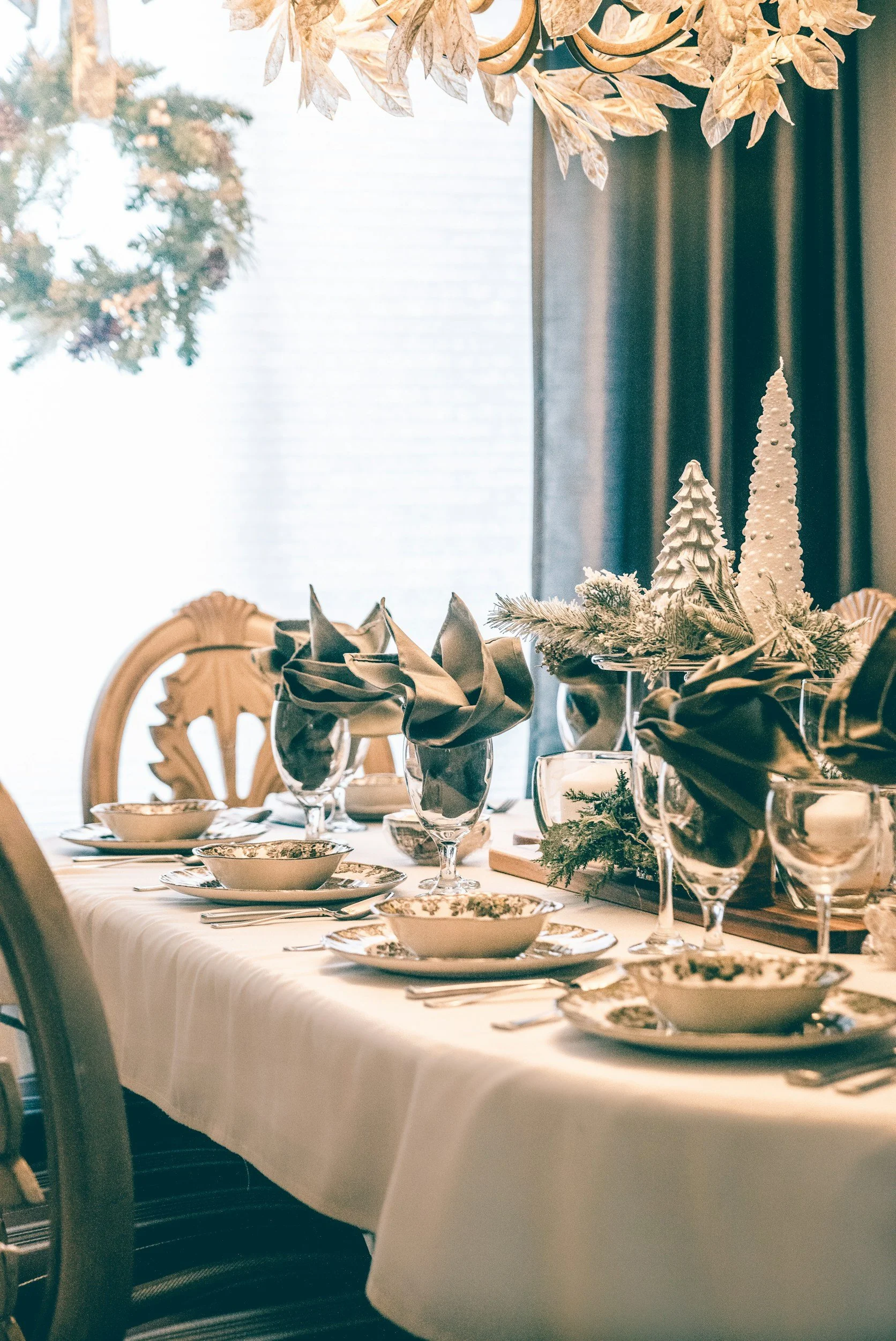 A festive dining table set for a holiday meal with fine china, glassware, and silverware. The table is decorated with gold and silver tableware, fabric napkins, and holiday-themed centerpieces that feature white Christmas trees and greenery. In the background, there is natural light coming through a window, a wooden chair, and a dark curtain.