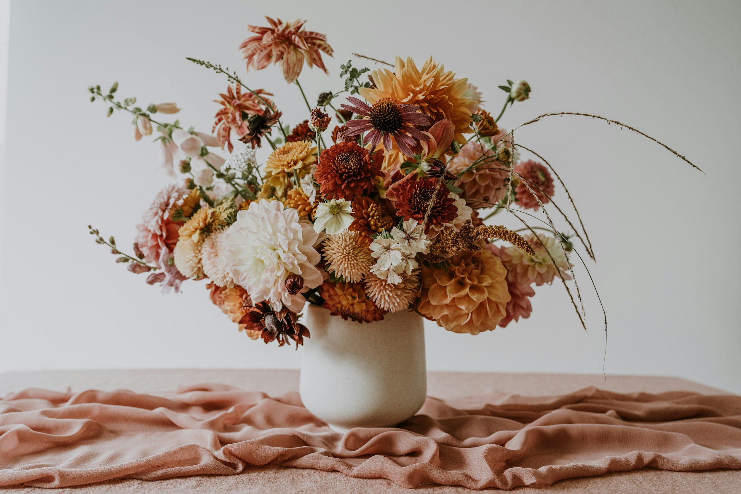 A bouquet of various flowers in shades of orange, peach, white, and burgundy in a white ceramic vase, placed on a pink satin cloth against a plain white background.