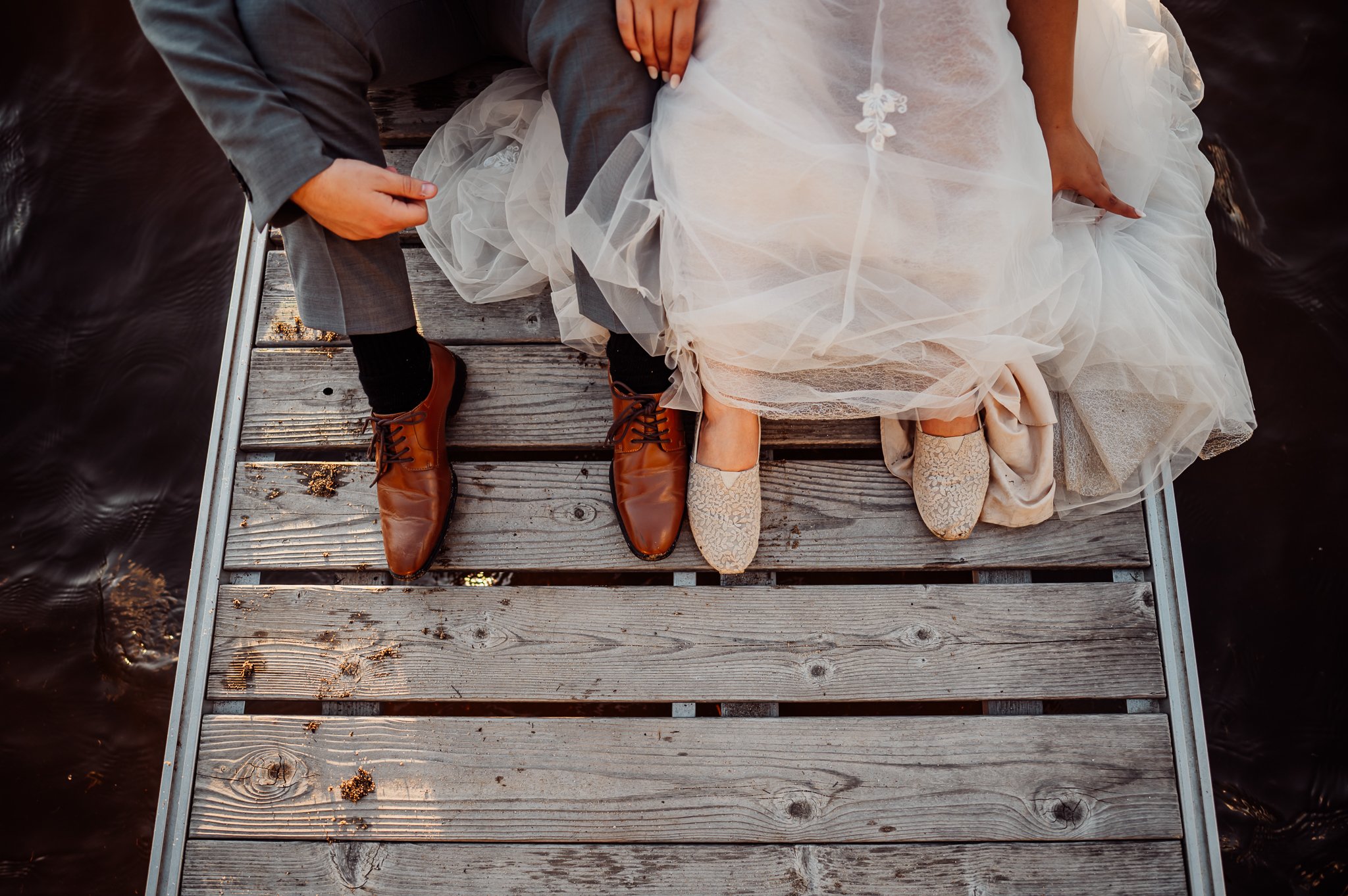 Wedding couple sitting on a dock, holding hands, with bride in a wedding dress and lace shoes, groom in gray suit, both wearing brown boots.