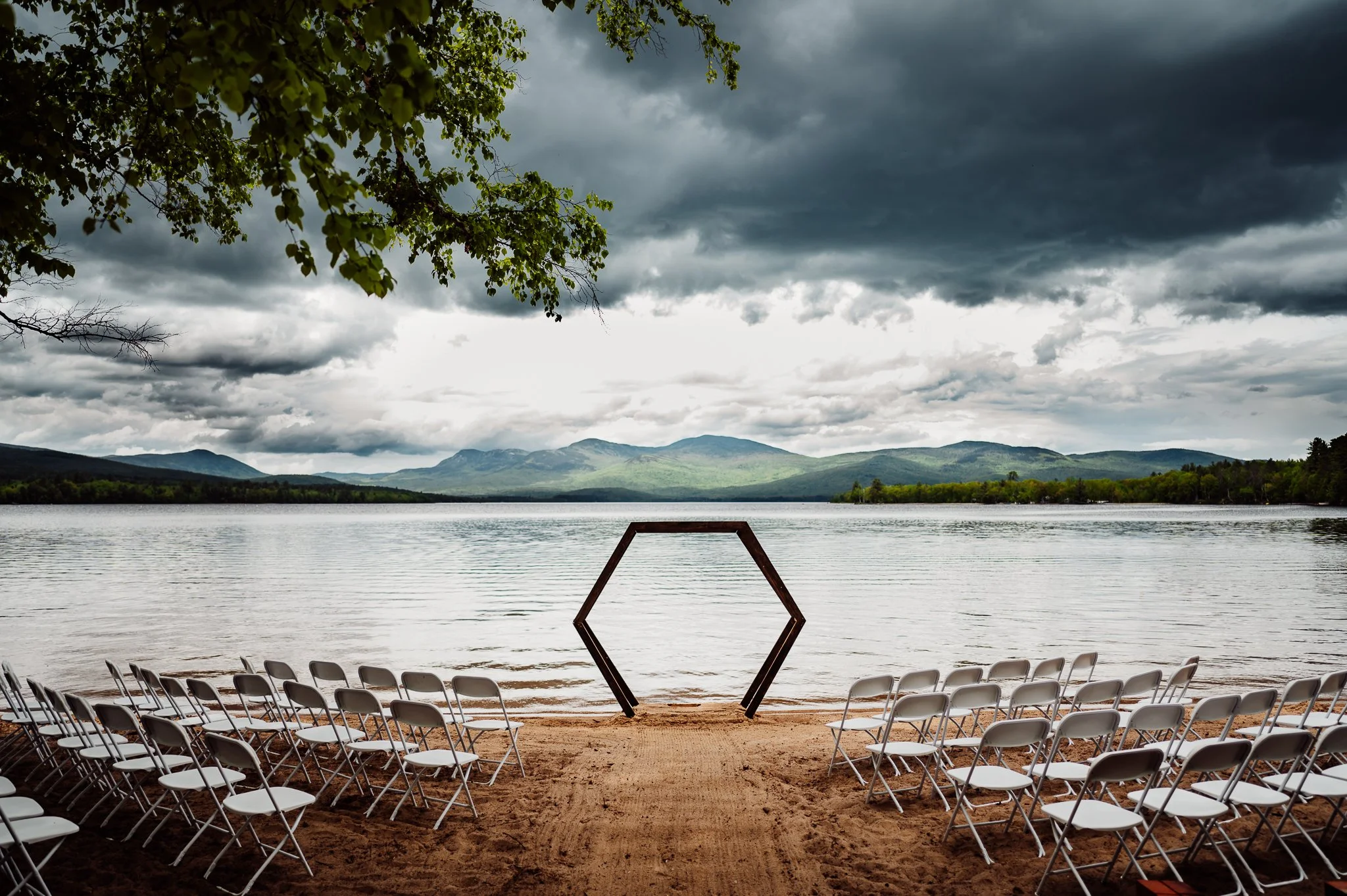 An outdoor lakeside setting with rows of white chairs arranged on sandy ground facing the water. A hexagonal metal frame stands in the center, suggesting a site for an event. The lake is calm, surrounded by green trees and distant mountains. Dark, stormy clouds fill the sky, indicating an approaching storm.