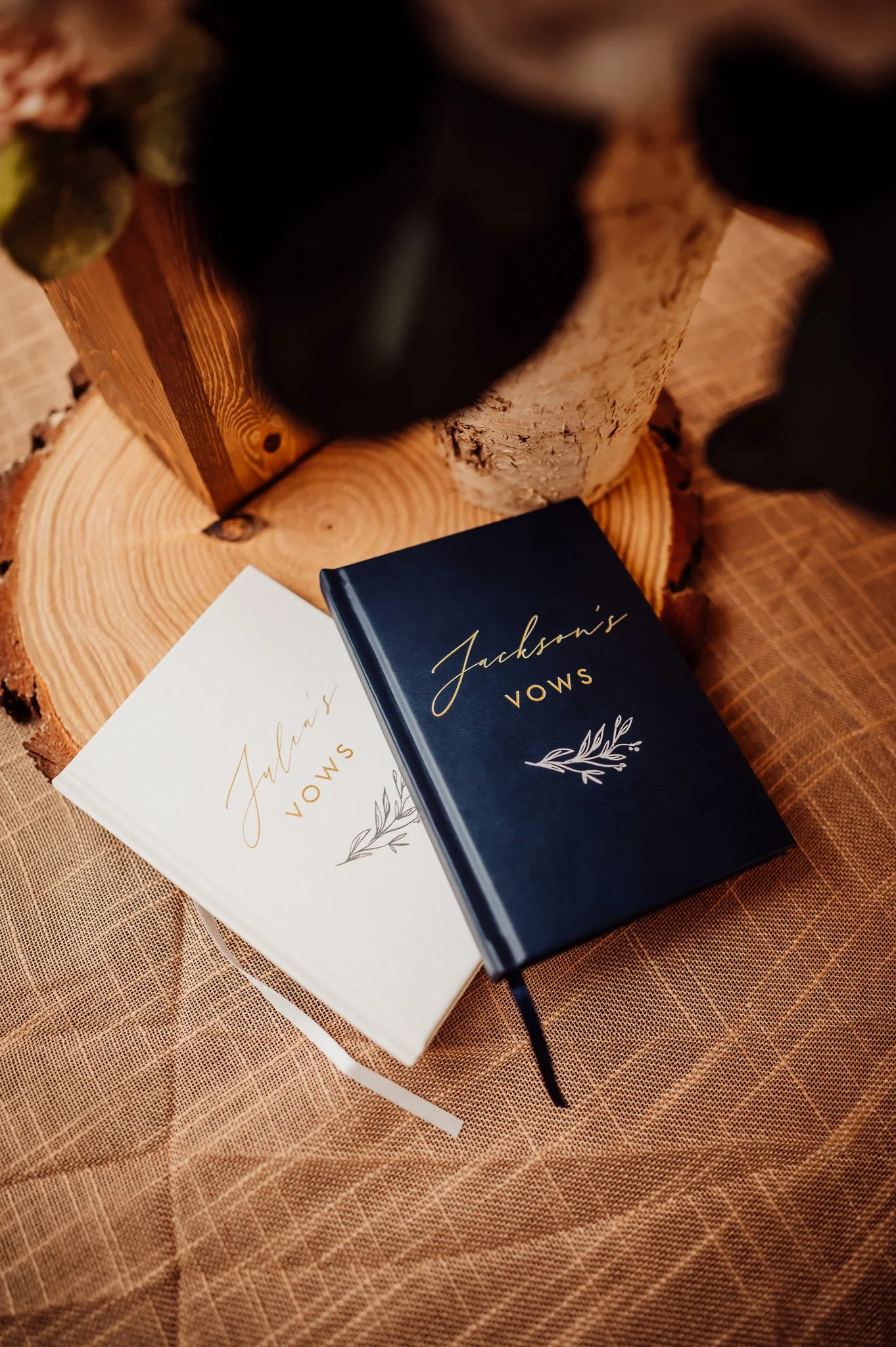 Two notebooks on a wooden log table, one white and one dark blue, with the title 'Jackson's Vows' and decorative foliage illustrations, placed on a textured fabric surface.
