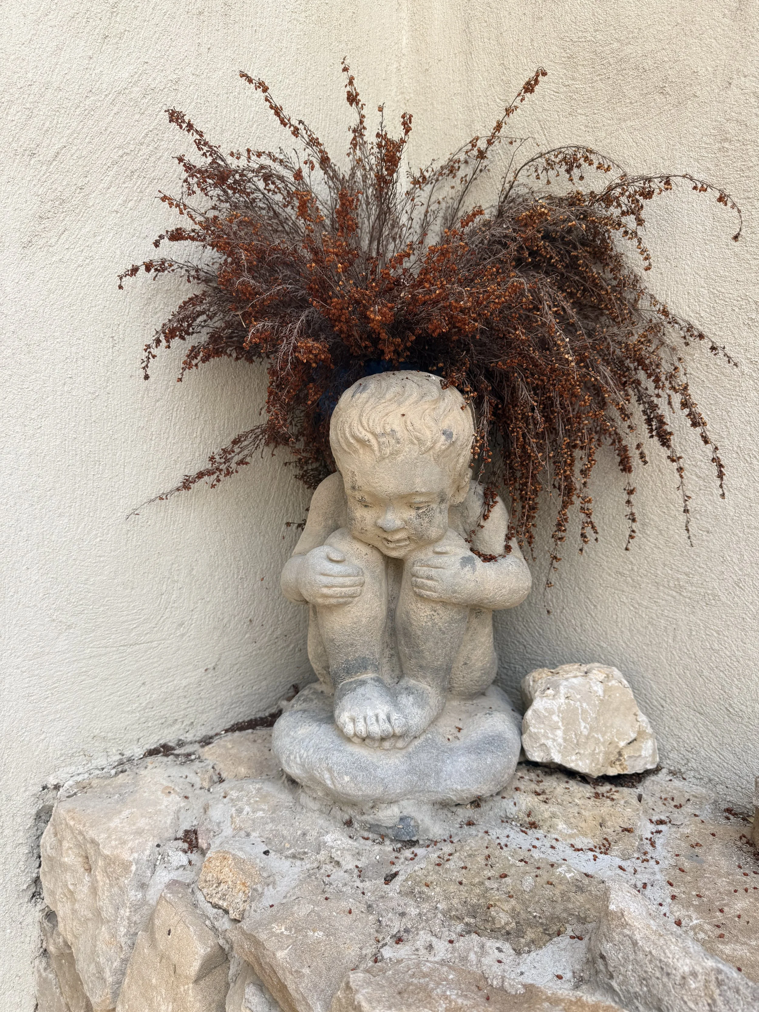 A stone sculpture of a child with closed eyes, holding its knees, situated on a rocky base against a beige textured wall. The sculpture has a rough surface and the child appears to be in contemplation or sorrow.