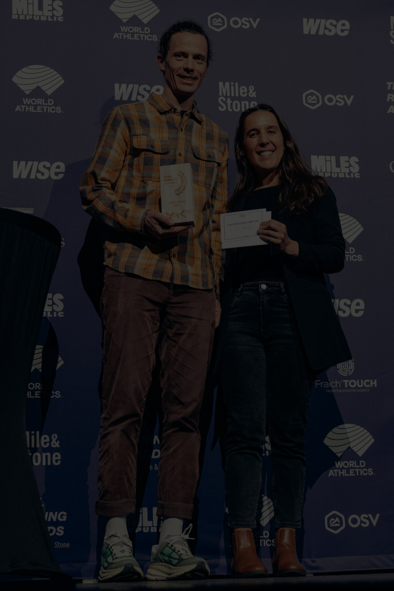 Deux personnes sur scène lors d'une cérémonie, une homme tenant un trophée et une femme tenant un chèque, devant un fond avec les logos des événements et des sponsors.