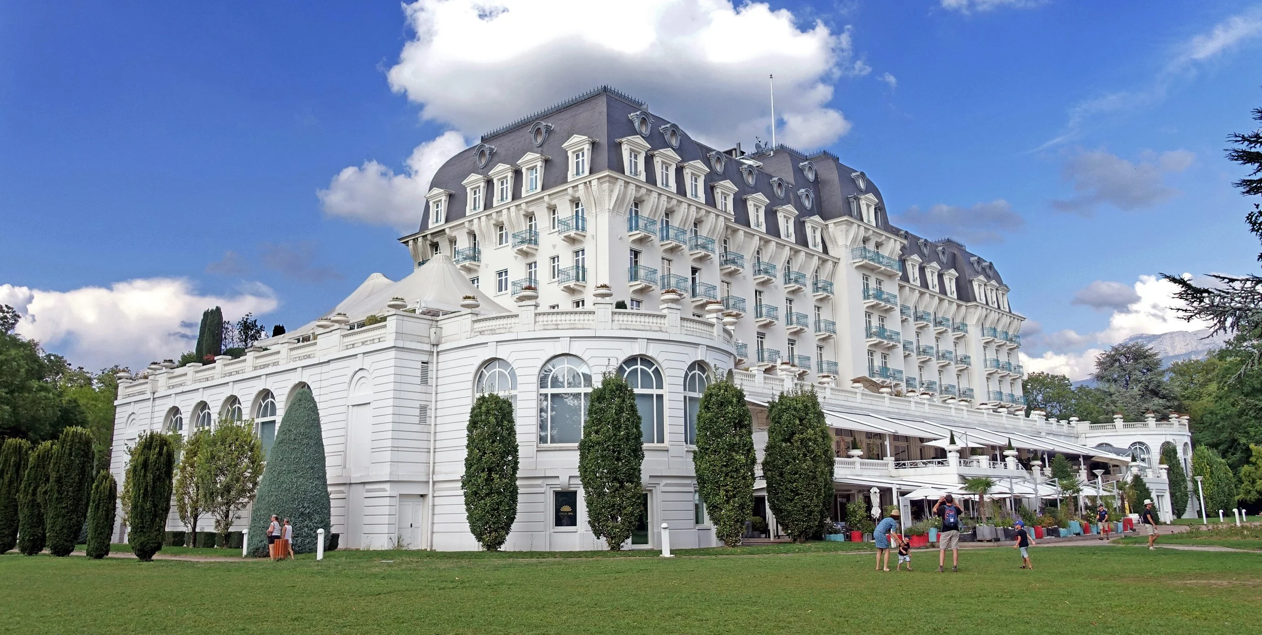 Un grand hôtel blanc avec plusieurs étages et balcons, entouré d'arbres et un espace vert où des gens sont assis ou jouent.