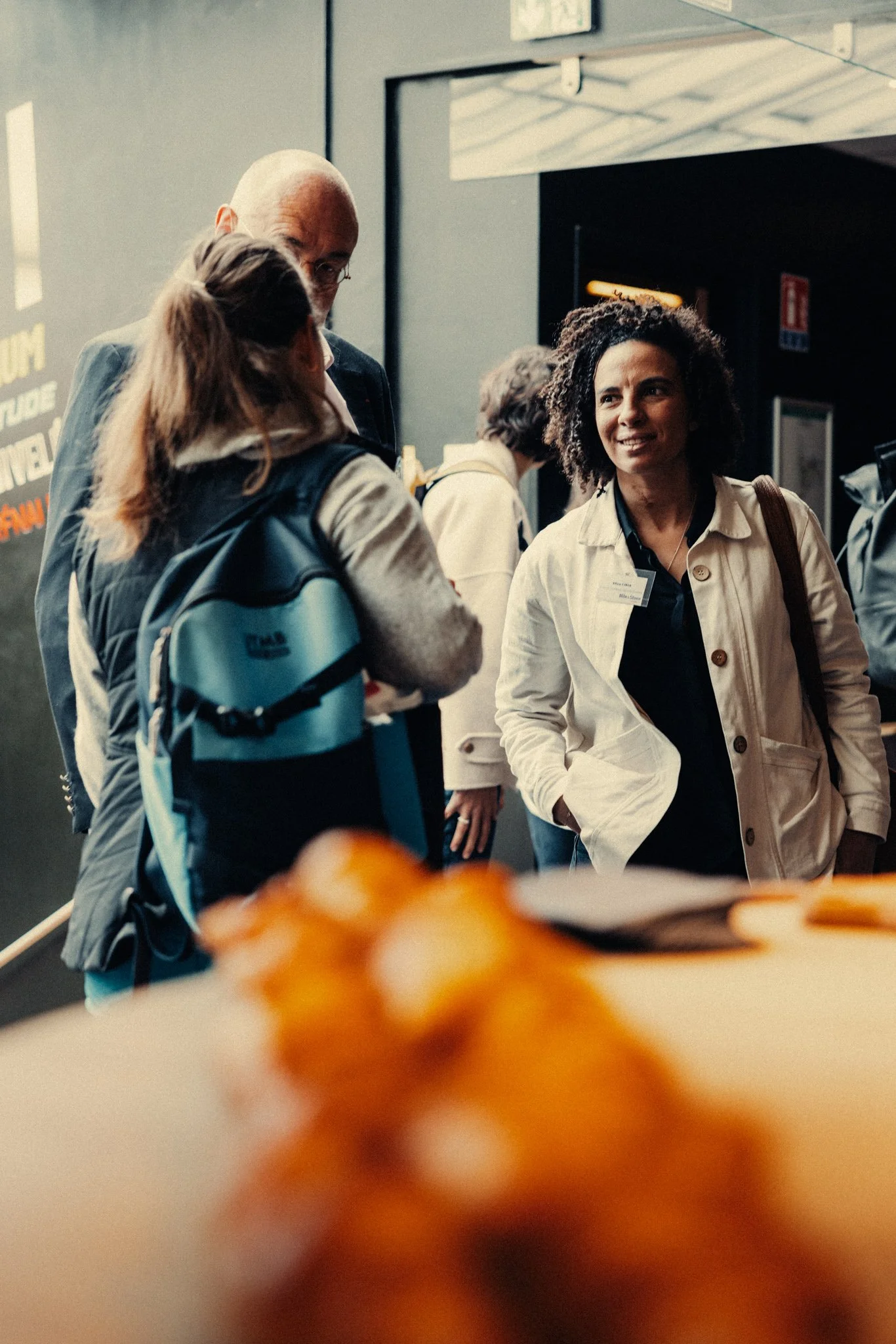 Groupe de personnes discutant dans un espace intérieur, une femme souriante portant une veste beige et une femme avec un sac à dos parlant avec un homme âgé. Température chaleureuse avec lumière naturelle.