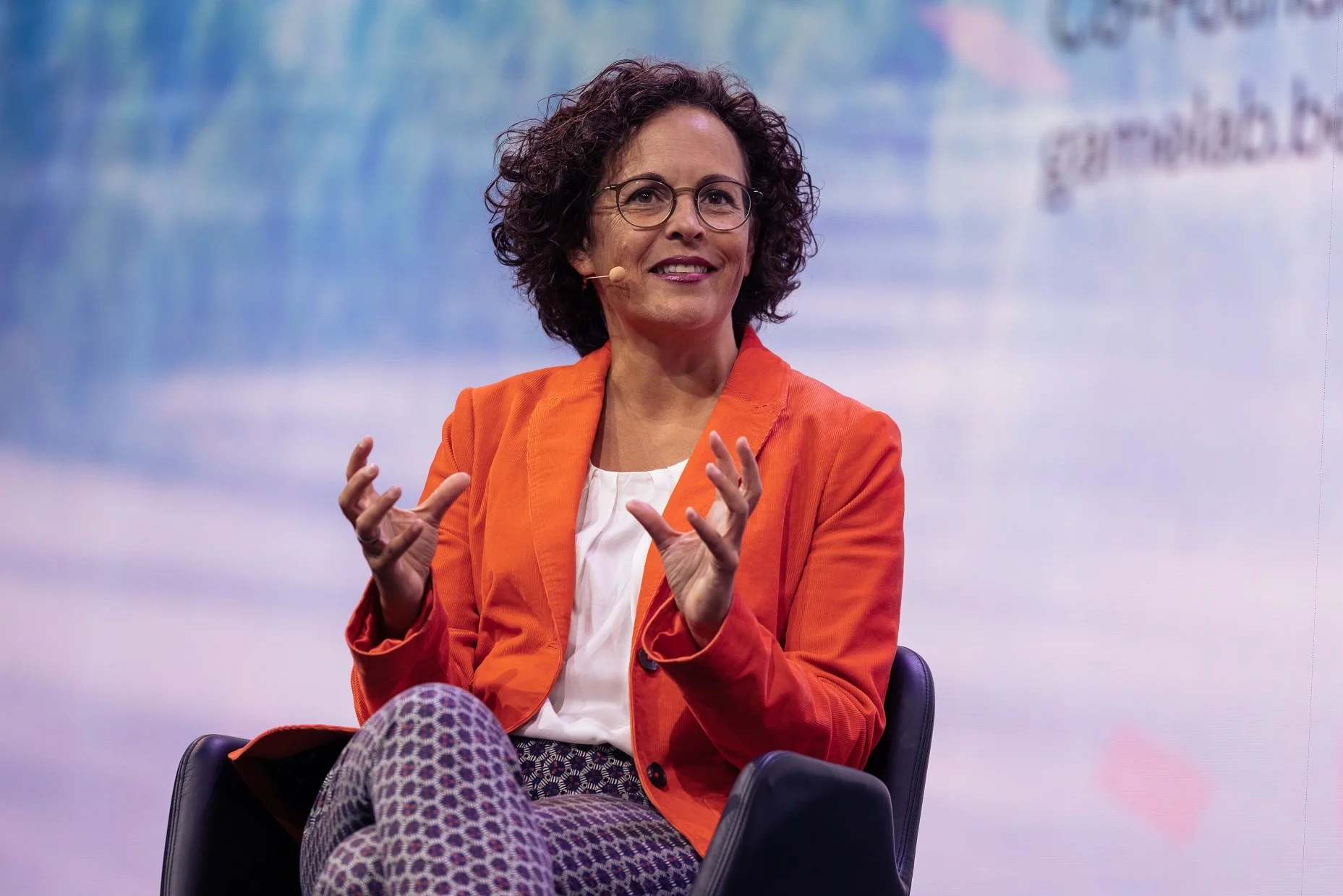 Nahaufnahme von Annette Wiedemann, Gründerin, von tbd - die Zukunftsberatung, mit Brille und einem orangefarbenen Blazer sitzt in einem Sessel auf einer Bühne, spricht, gestikuliert mit den Händen; Strategiekommunikation, Storytelling