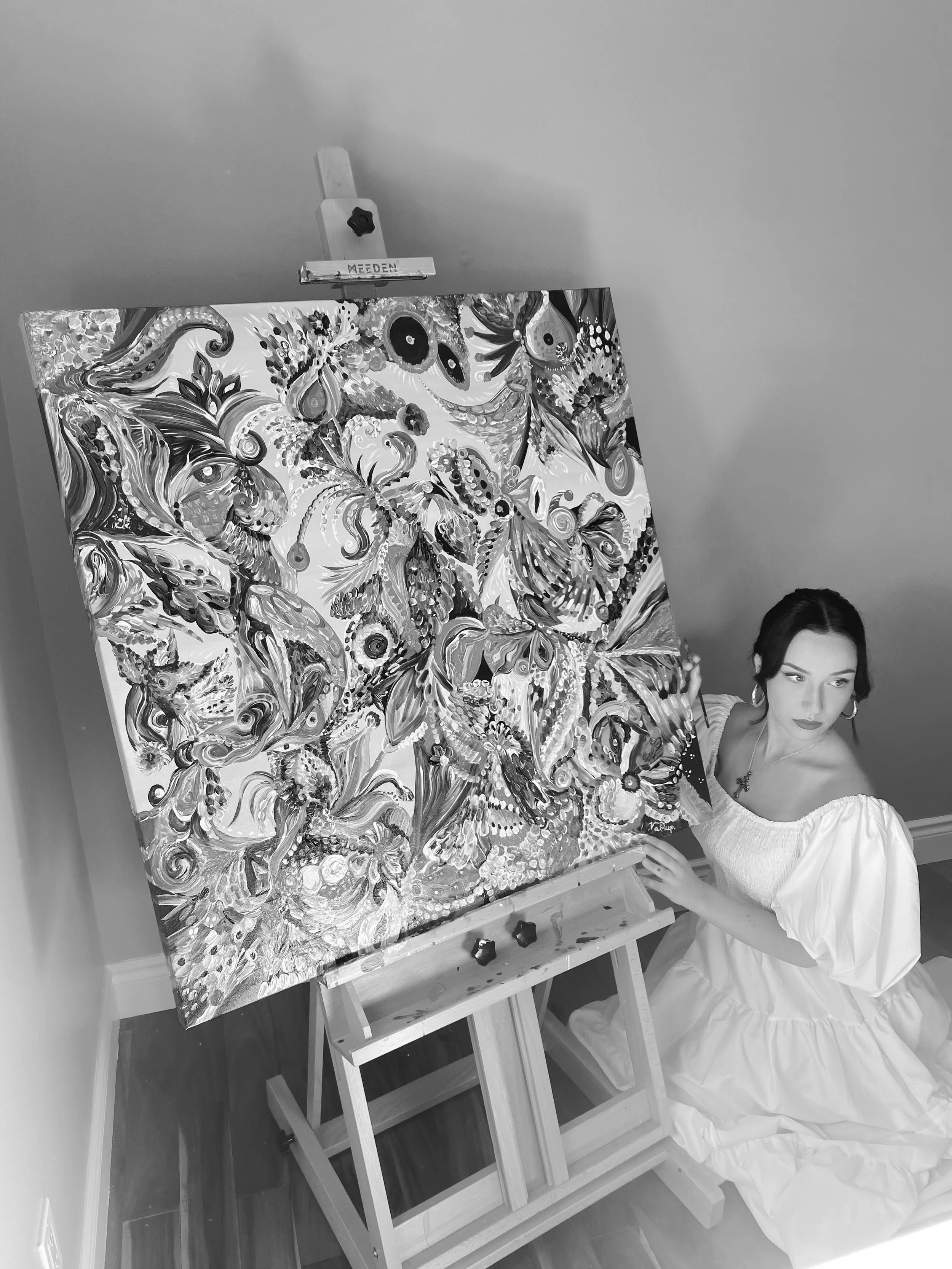 A woman in a white dress is kneeling next to an easel, holding a paintbrush, and working on a large, detailed, and colorful painting that features floral and decorative elements.