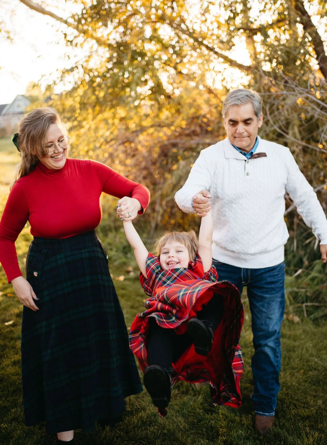 Family of three, a woman, a man, and a young girl, outdoors during autumn. The girl is sitting in a red plaid stroller, smiling and wearing a matching red plaid dress. The woman and man are holding her hands and swinging her in the air, all smiling. The background features trees with fall-colored leaves.