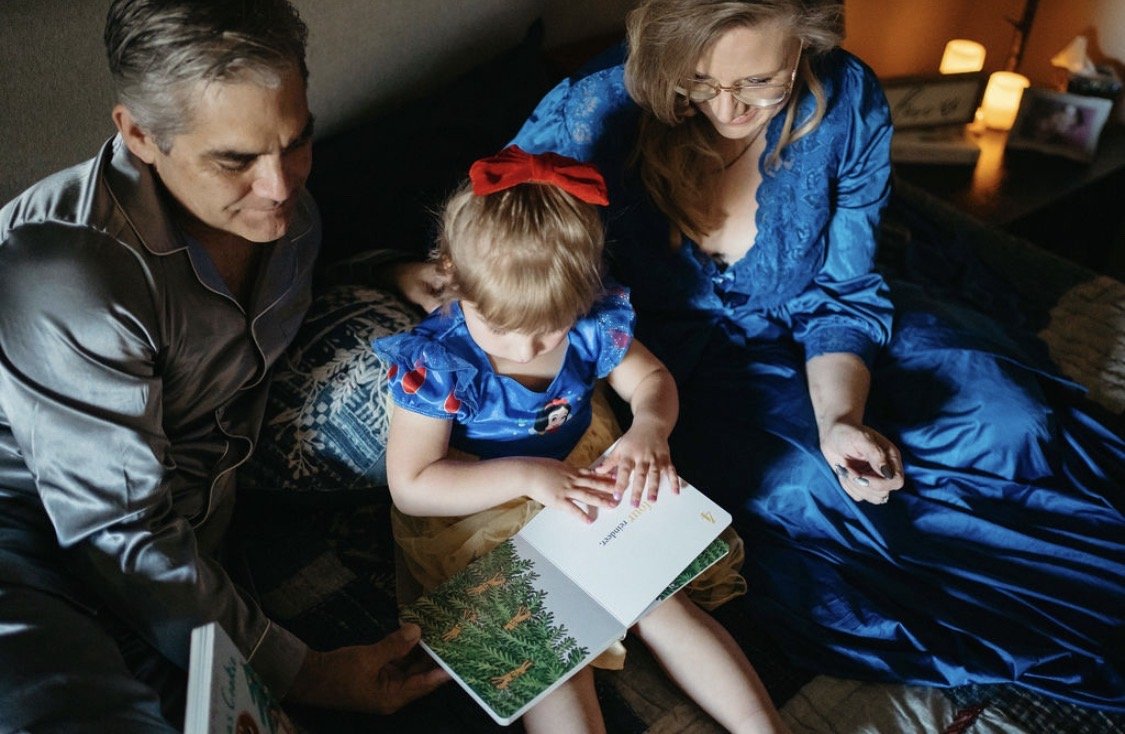 A family of three sitting on a bed together, reading a children's book with Christmas-themed illustrations.