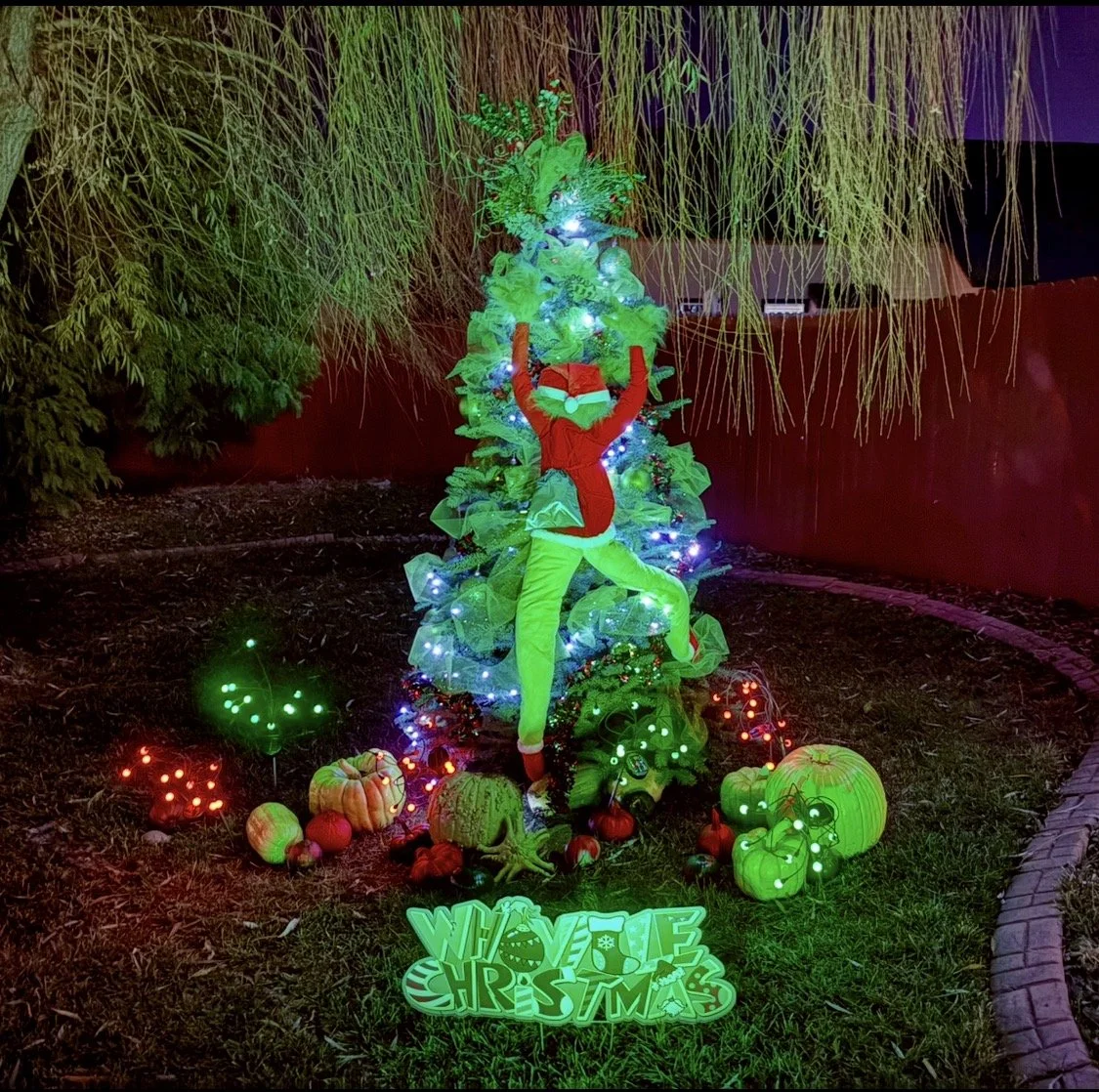 Christmas display with a decorated tree, a woman dressed as an elf, and pumpkins, lights, and a sign reading 'WOLVE CHRISTMAS' at night.