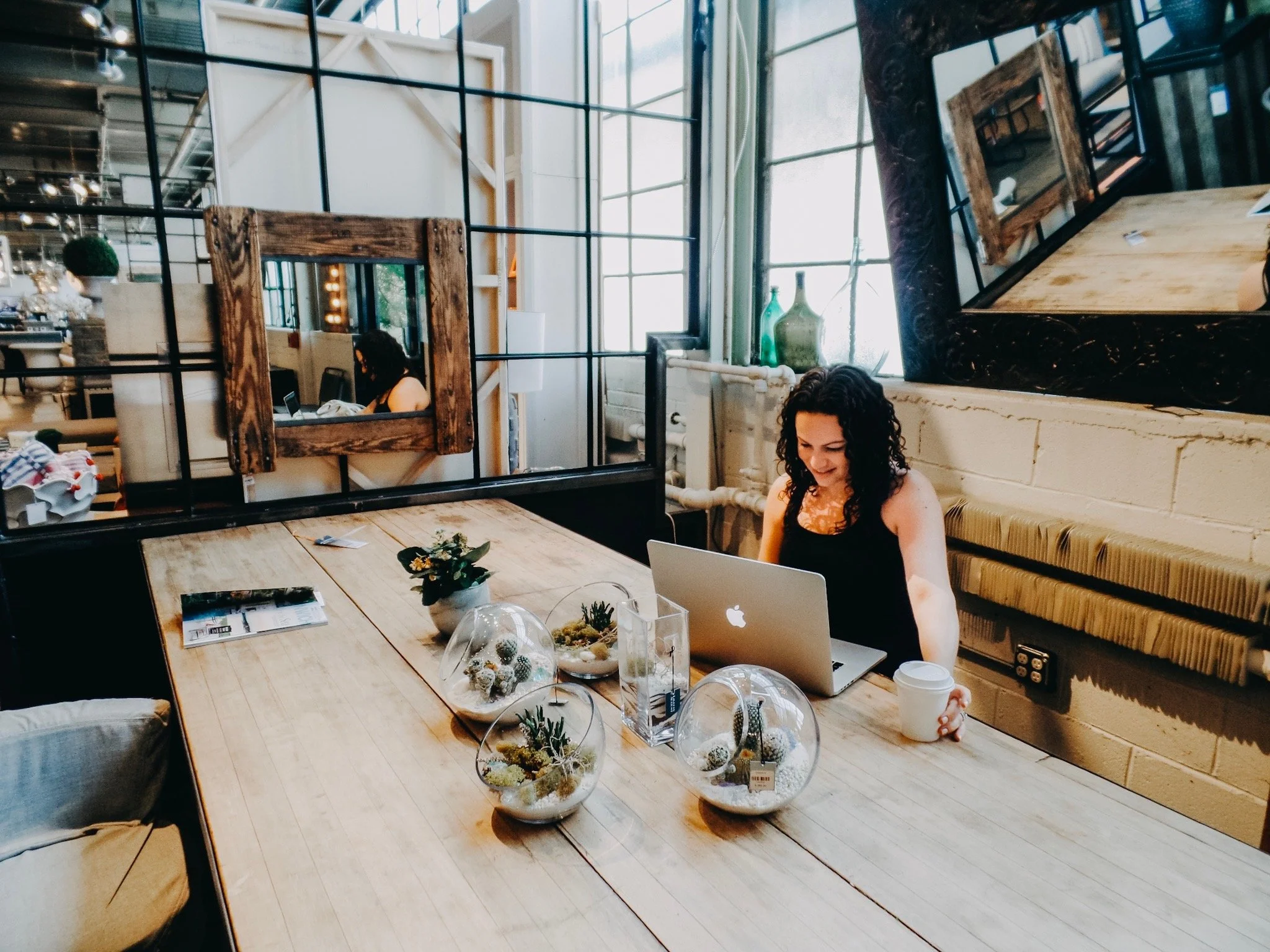 Interior of a modern cafe or restaurant with large windows, a long wooden table decorated with plants and decorative objects, and a woman working on a laptop. Mirrors and industrial-style decor are visible.