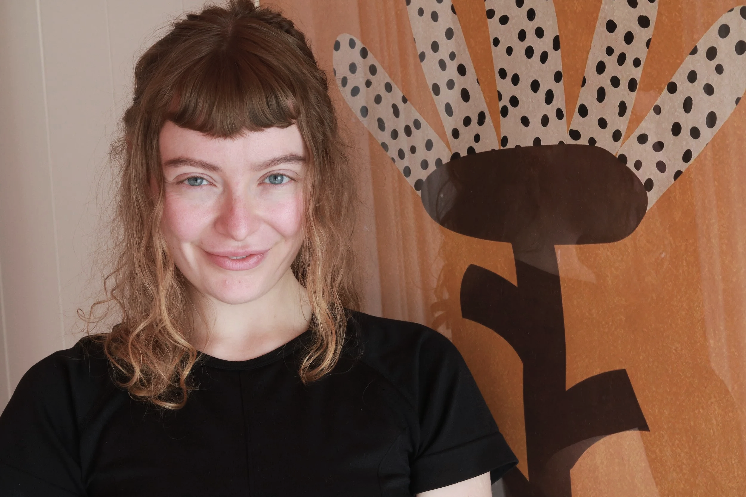 A young woman with curly hair and light skin in front of a wall with bold orange and black graphic art.