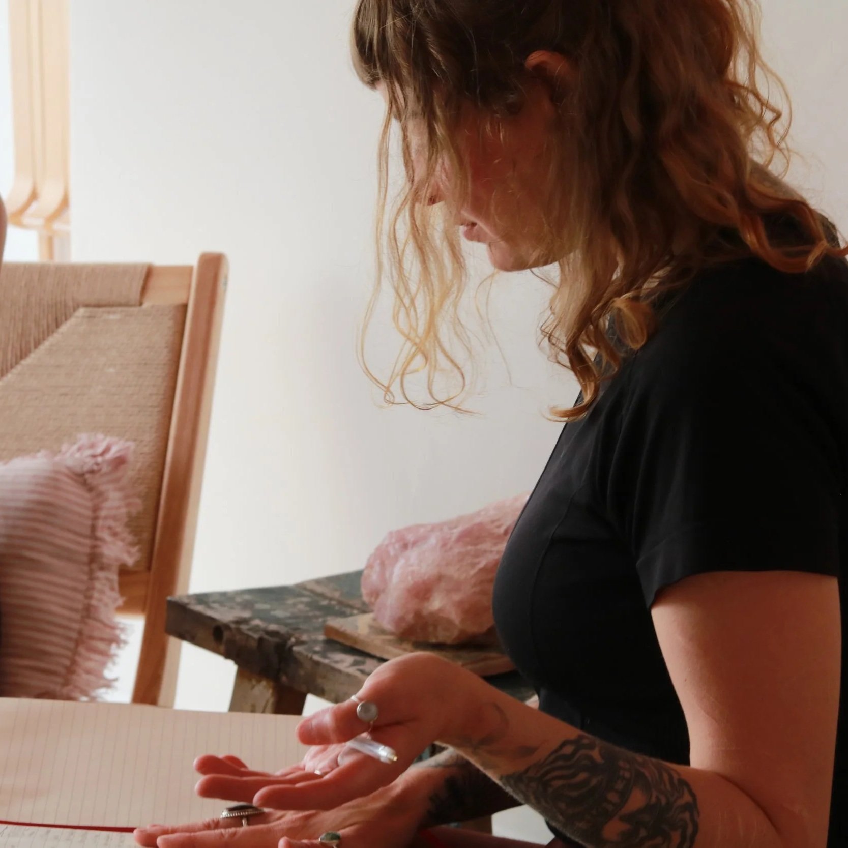 A woman with curly red hair writing in a notebook on a table, wearing a black shirt, with a partially visible tattoo on her arm.
