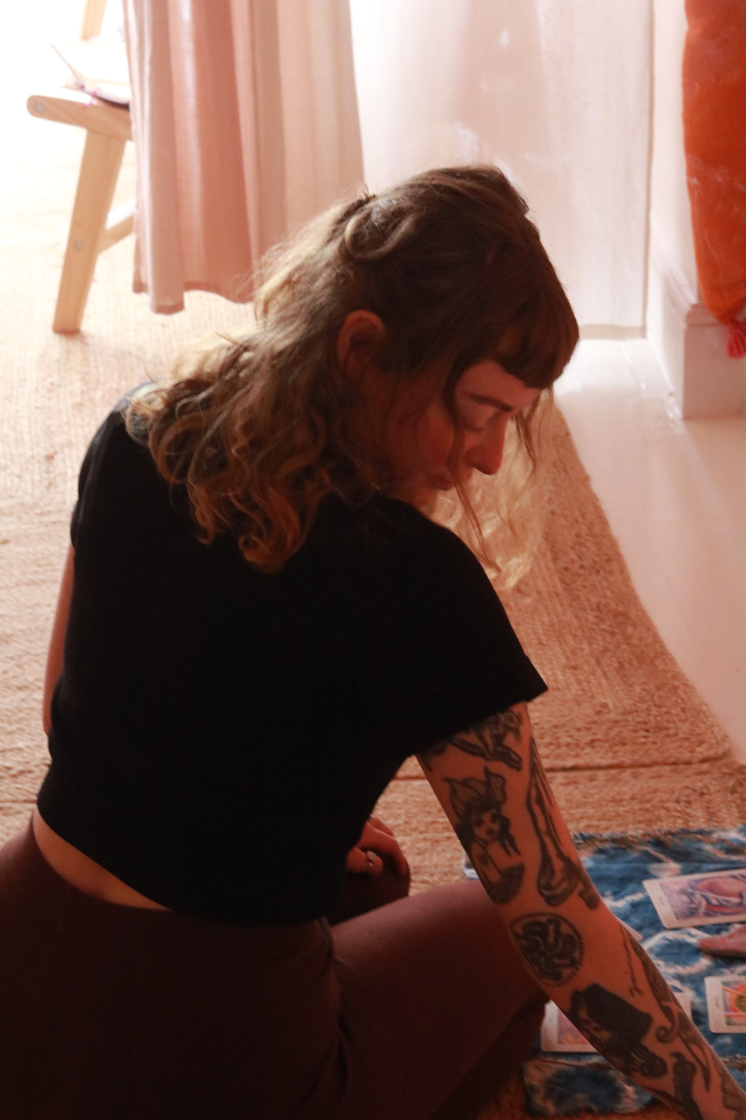 A person with curly, shoulder-length hair and tattoos on their arms, sitting on the floor next to a bed with a quilt or blanket. The person is looking downward and is wearing a black shirt.