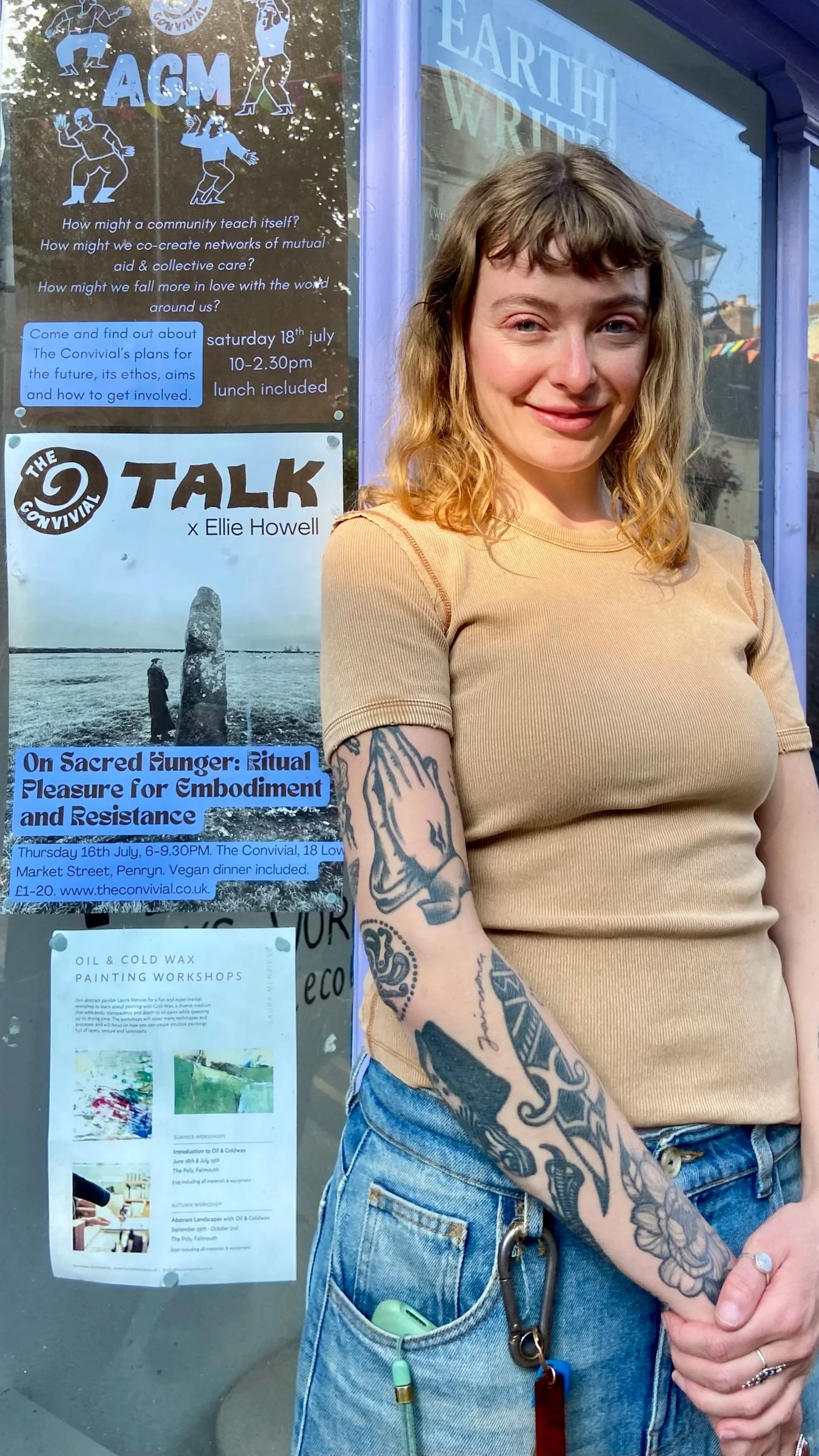 A young woman with tattoos on her arms, wearing a tan short-sleeved shirt and blue jeans, standing in front of a glass display board with various posters, including one promoting an event 'The Convivial' and another about oil and cold wax painting workshops. She has curly hair and is smiling slightly.