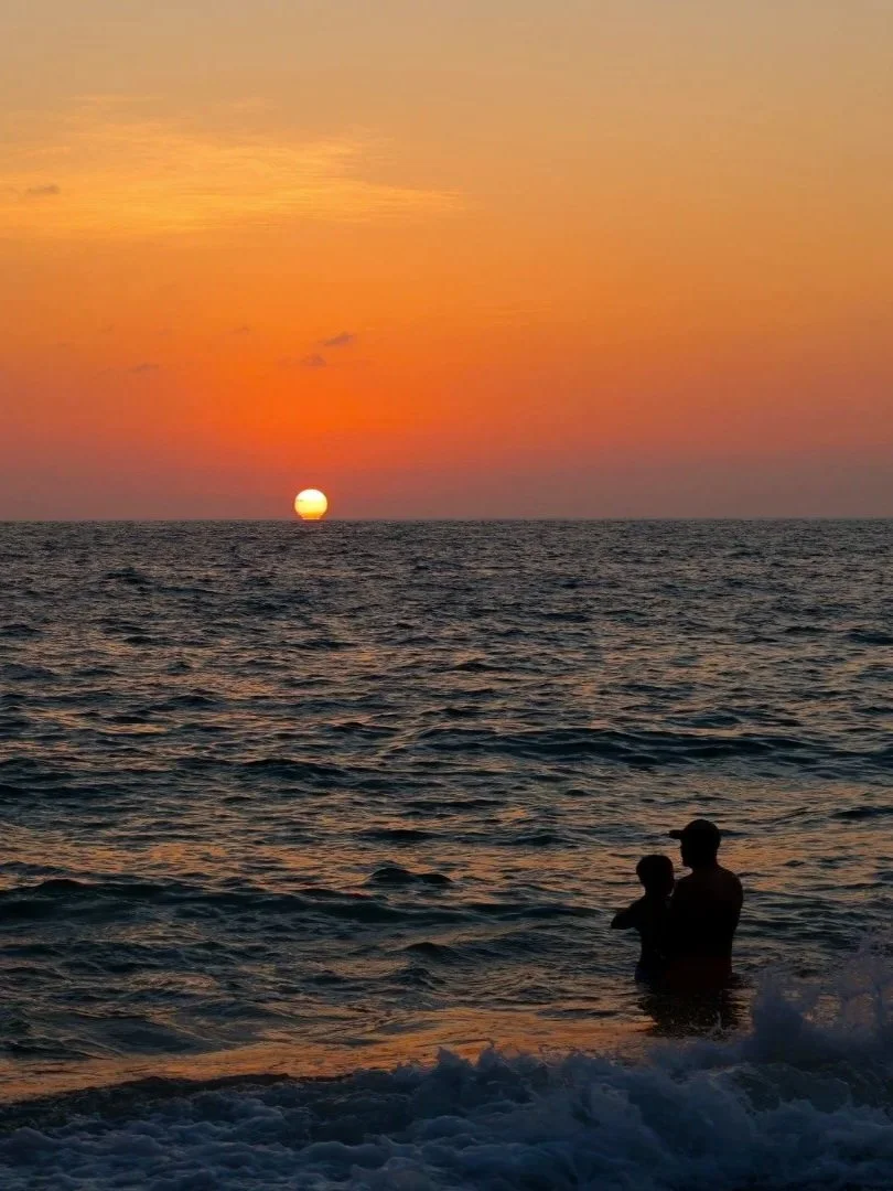 Silhouette of a person holding a child in the ocean during sunset, with orange and pink sky and the sun near the horizon.