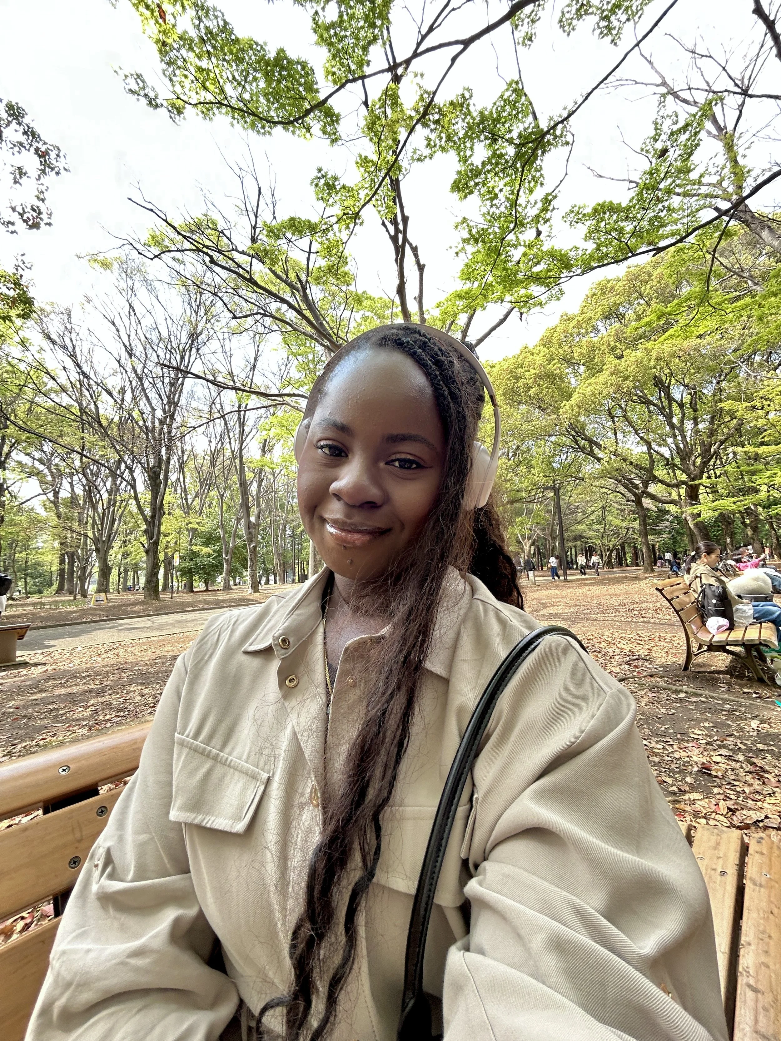 A young woman with long dark hair wearing headphones and a beige jacket, smiling while sitting on a park bench surrounded by trees with green and yellow leaves, and other park visitors in the background.