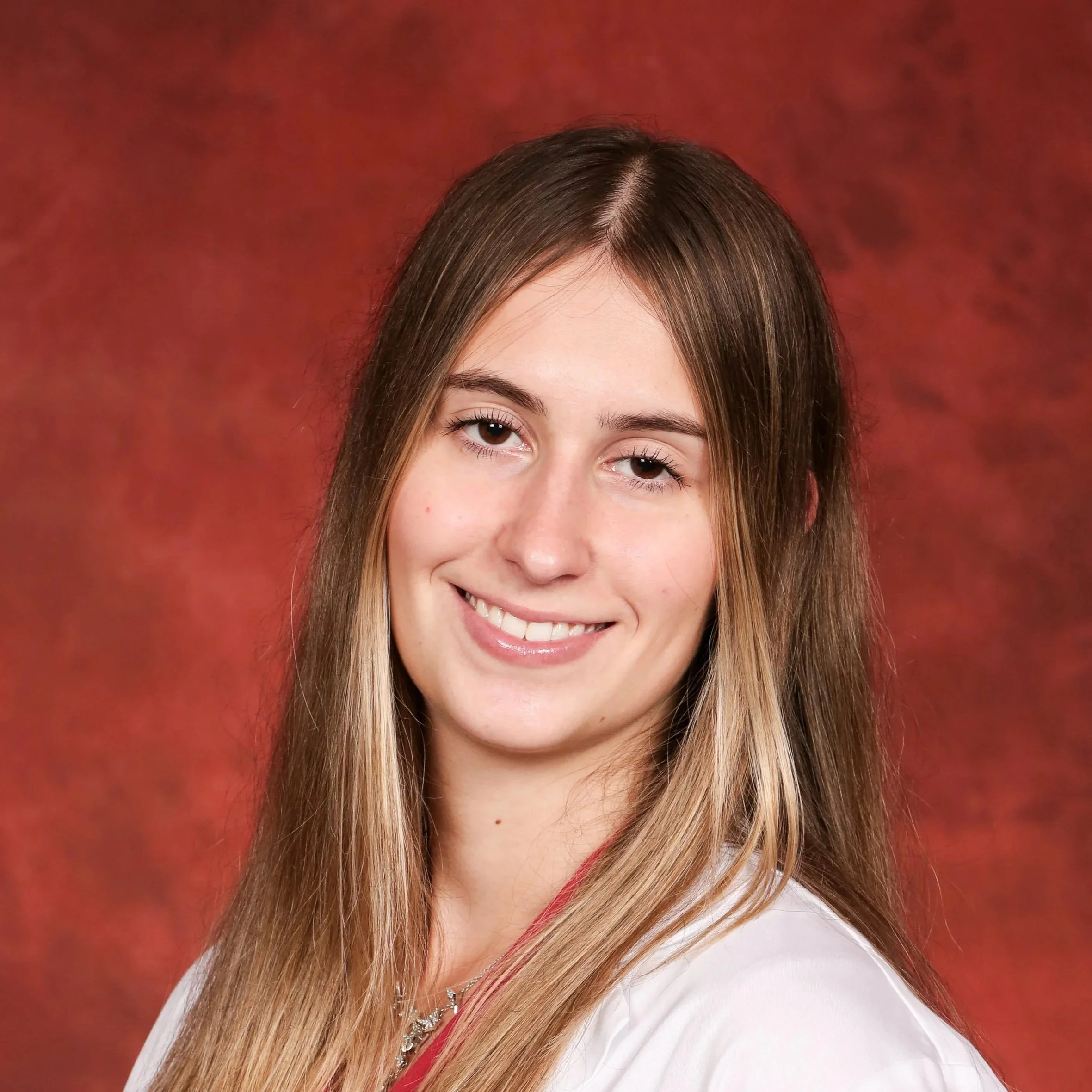 Close-up of a woman with long, straight blonde hair smiling at the camera against a red patterned background, wearing a white top and a necklace.