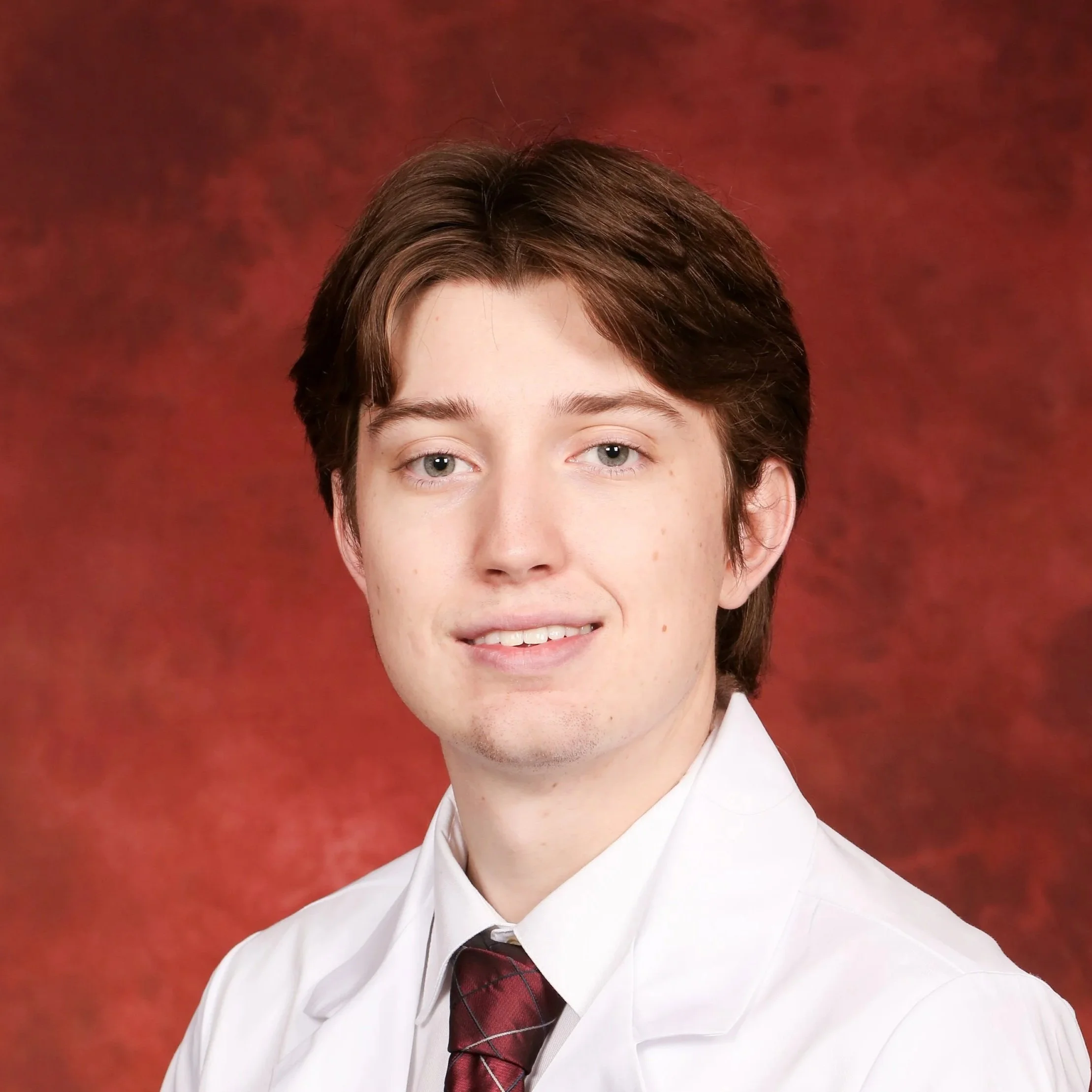 Close-up portrait of a young person with short brown hair and blue eyes, wearing a white dress shirt and a dark red and black plaid tie, against a red mottled background.
