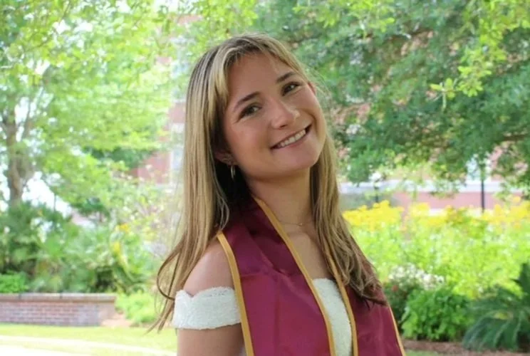Young woman smiling outdoors wearing a graduation stole, surrounded by green trees and foliage.