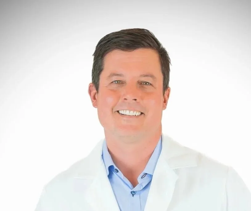 A professional man wearing a white lab coat and a blue collared shirt, smiling in front of a plain white background.