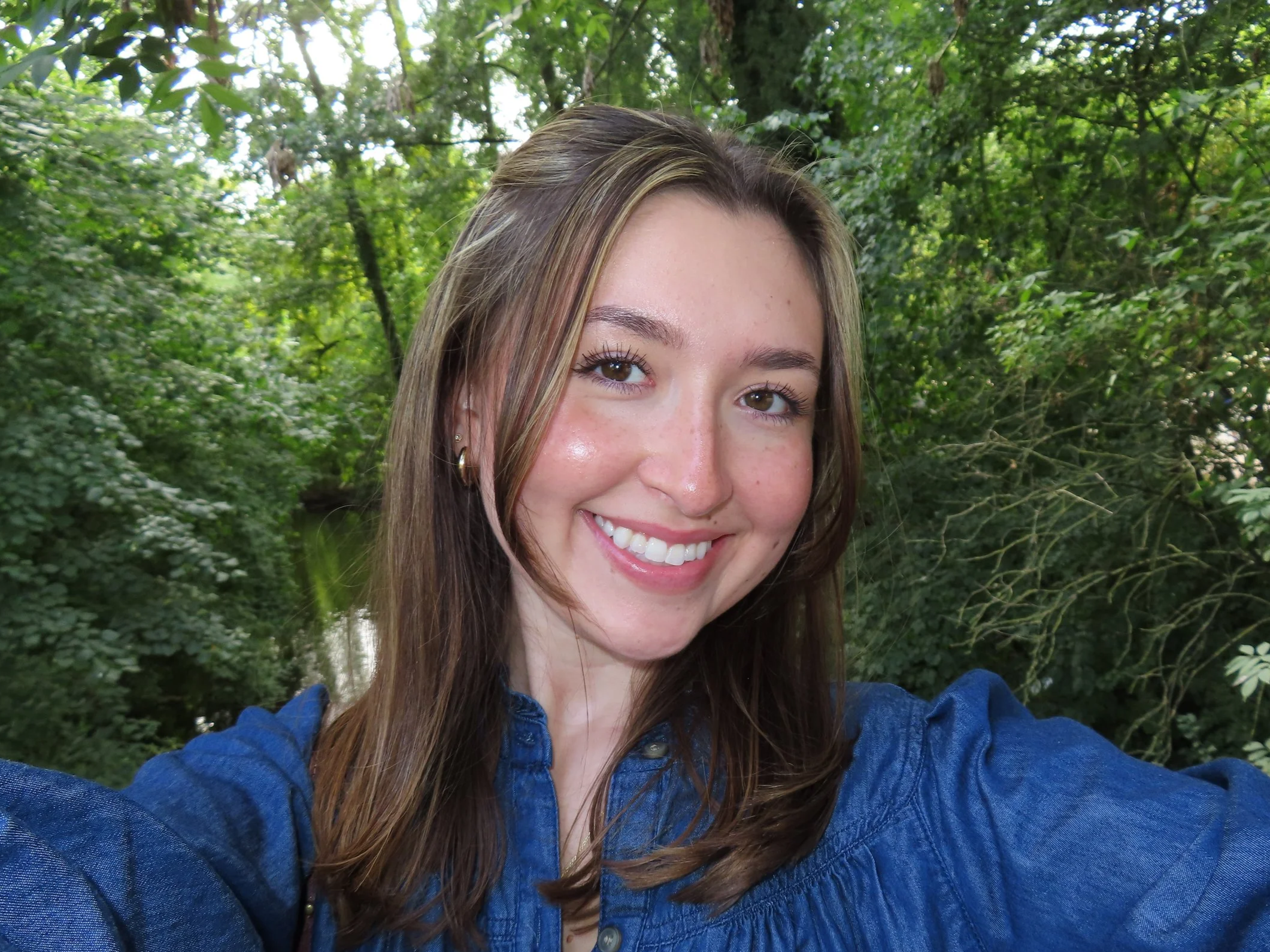 A young woman with long brown hair and freckles smiling, taking a selfie outdoors in front of green leafy trees.