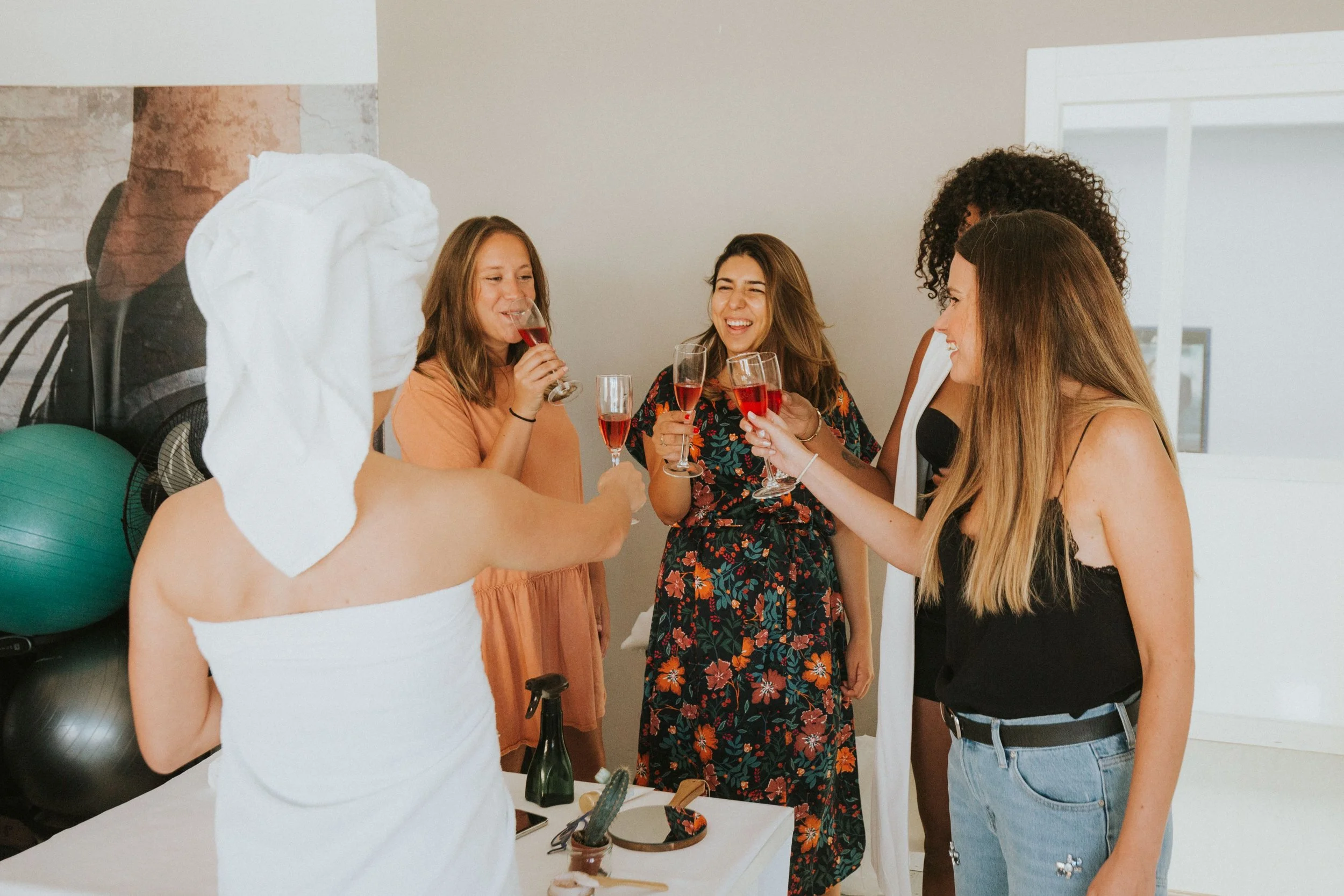 Group of five women smiling and holding glasses of rosé wine, celebrating in a bright indoor space with a white wall and large window.