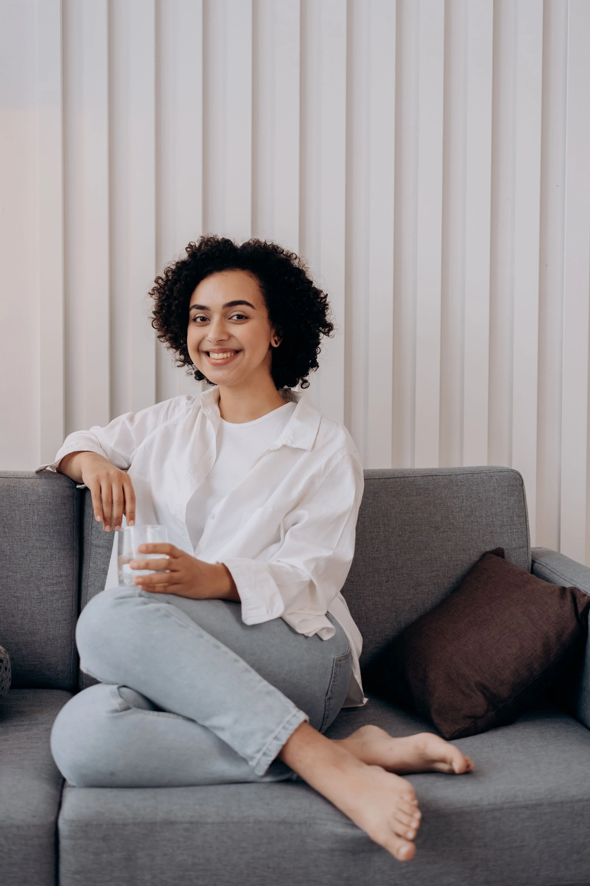 Woman with curly hair sitting on a gray couch, smiling, holding a glass of water, wearing a white shirt and light jeans, in a room with a cream wall and dark brown cushions.