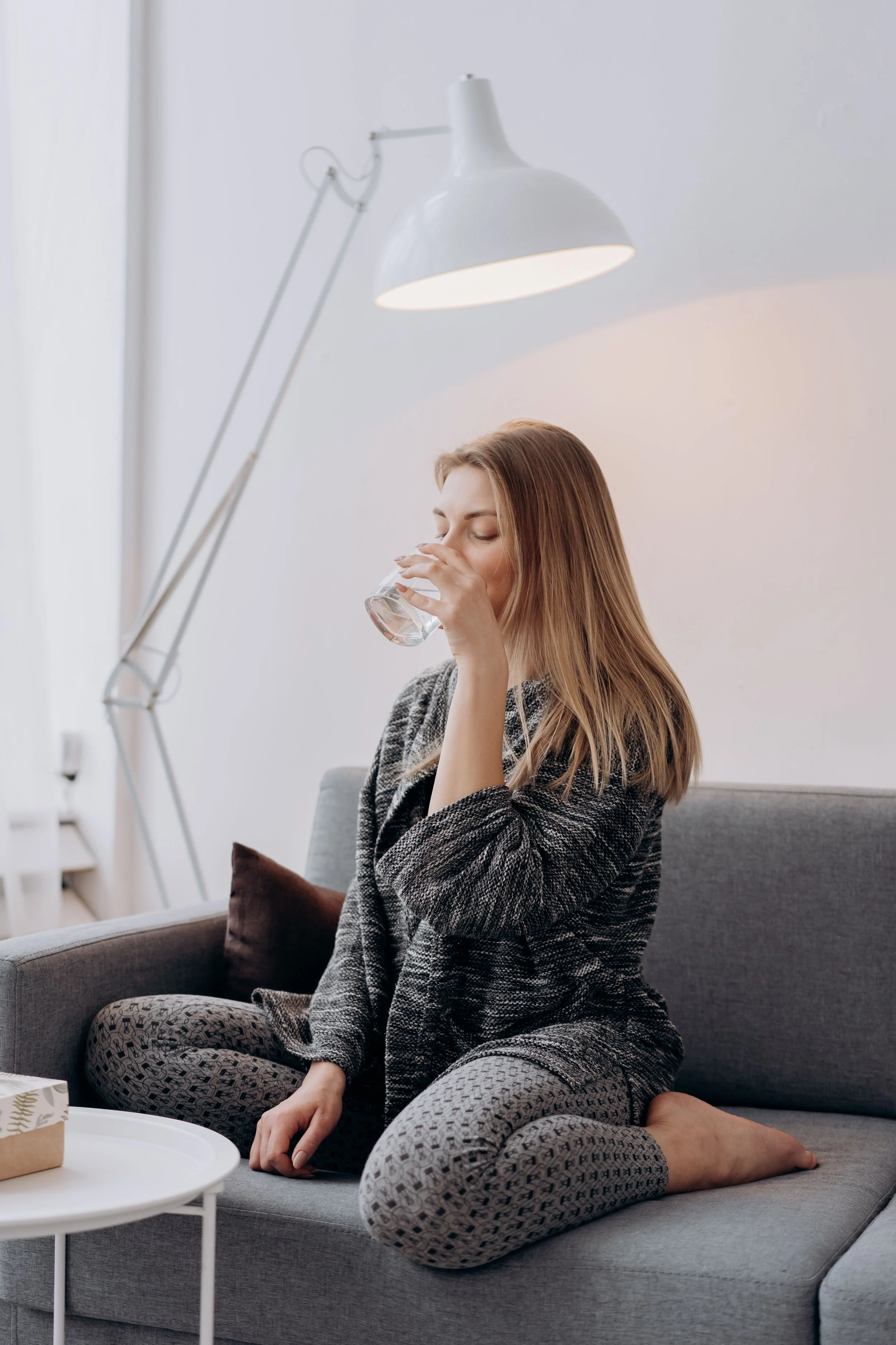 A young woman with long blond hair, wearing pajamas and a cozy robe, is sitting on a gray sofa, drinking water from a glass, with her eyes closed in a relaxed manner.