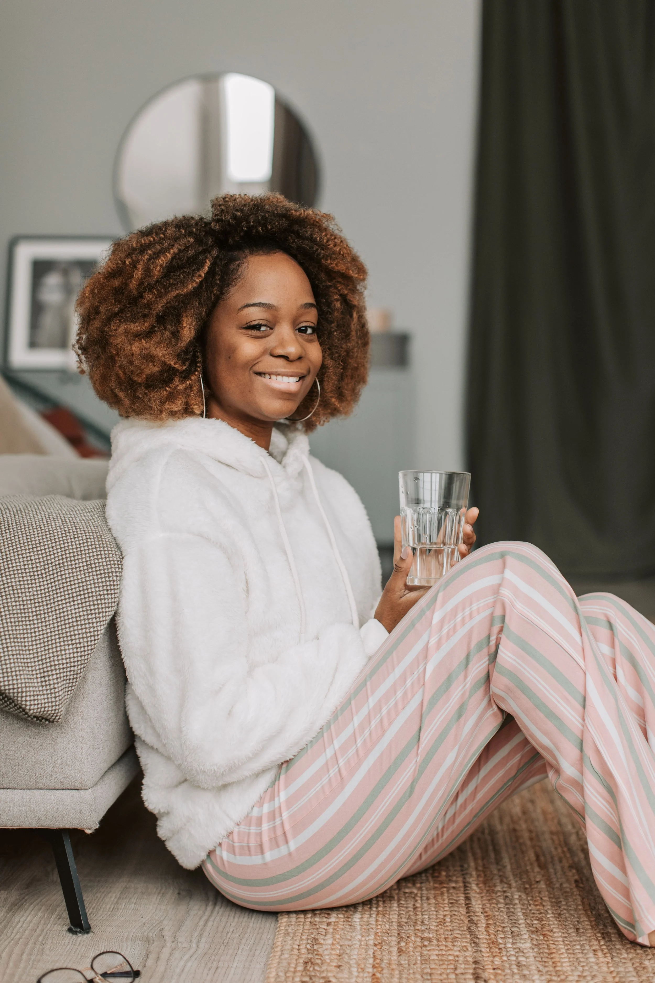 A young woman with curly hair, wearing a white hoodie and striped pajamas, sitting on the floor next to a sofa, holding a glass of water, smiling at the camera.