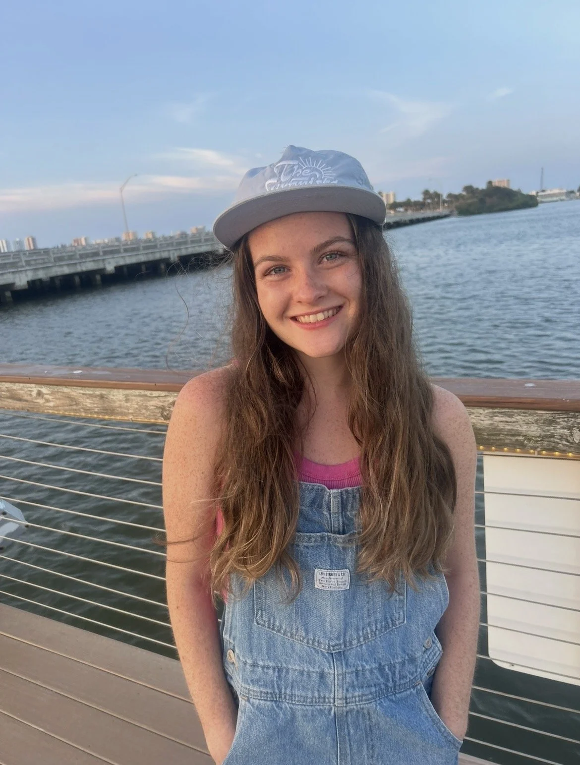 A young woman with long wavy brown hair smiling outdoors near a body of water, wearing a gray cap and denim overalls, during the daytime.