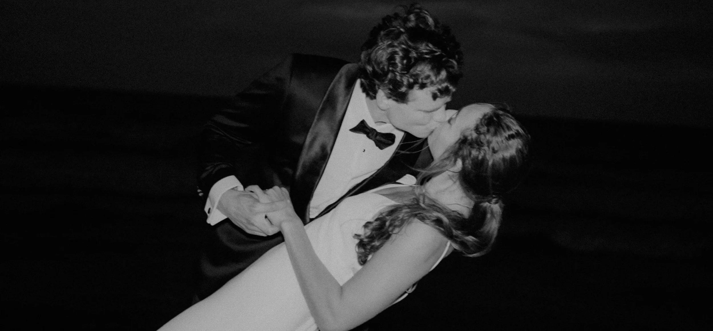 Black and white photo of a young couple in formal attire, sharing a kiss and holding hands, on a dark background.