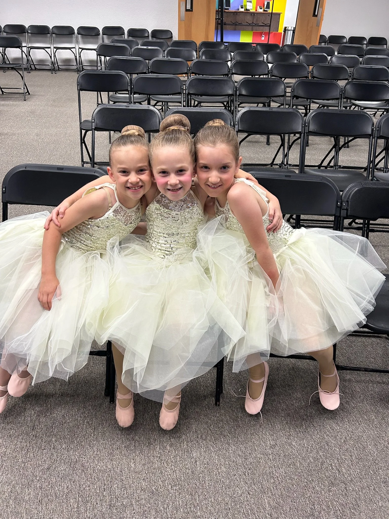 Three young girls dressed in ballet costumes sitting closely together in an auditorium with rows of empty black chairs behind them.