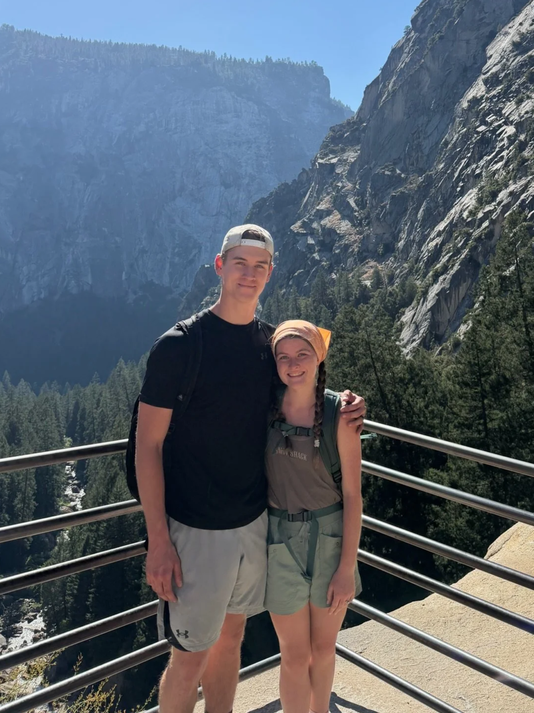 A young man and woman standing on a viewing platform with a mountain and forested landscape in the background.