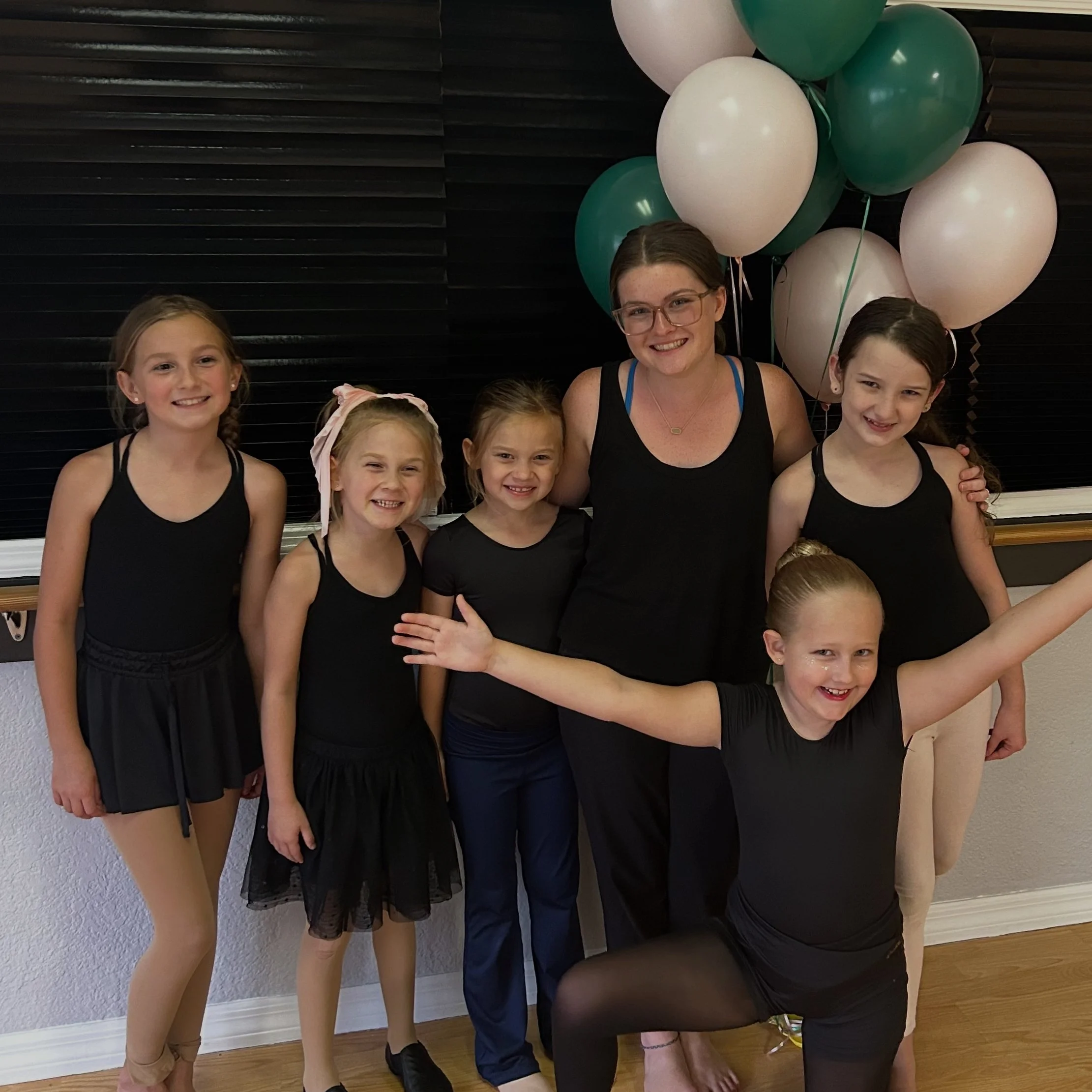Group of young girls and an adult woman dressed in black at a party or celebration, standing in front of a black wall with green and white balloons.