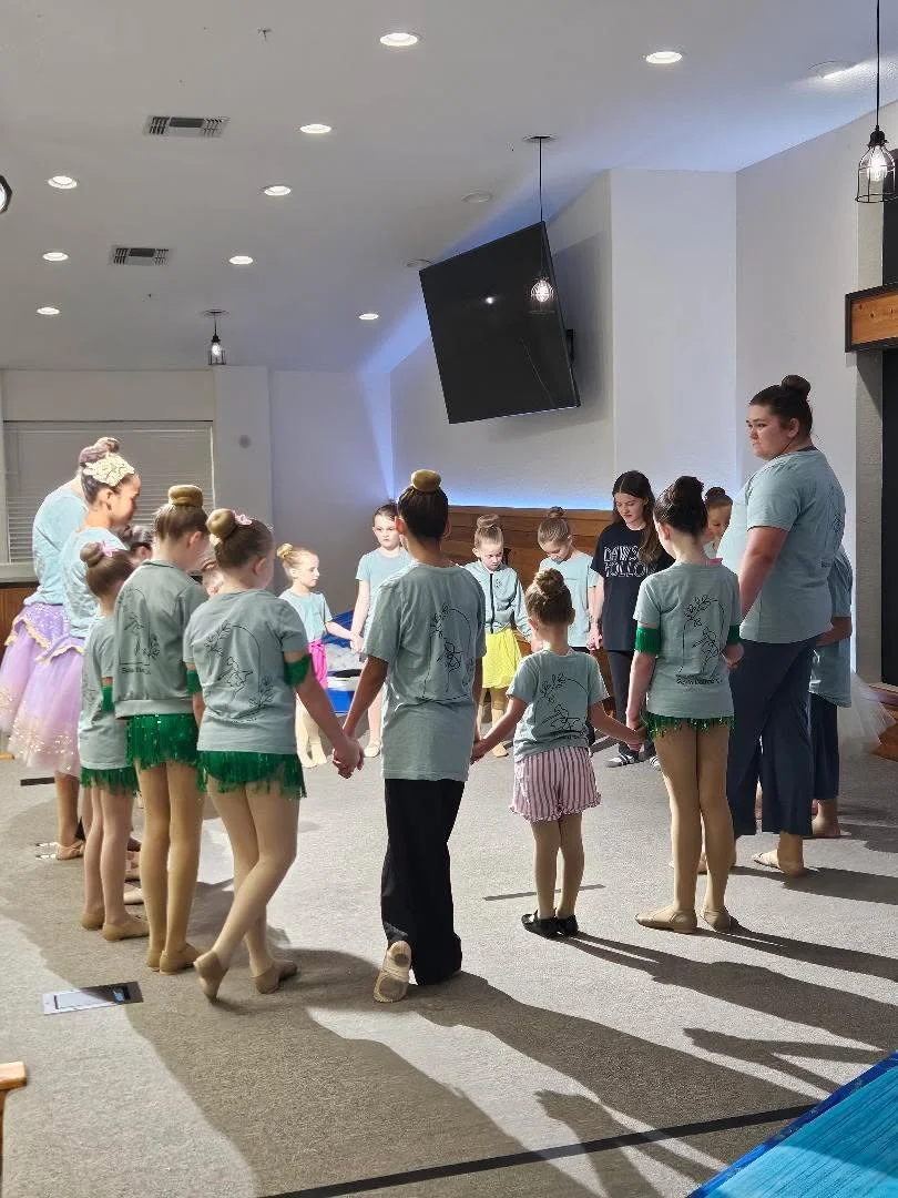 Children and instructors holding hands in a circle during a dance or ballet practice in a room with a large monitor and bright ceiling lights.